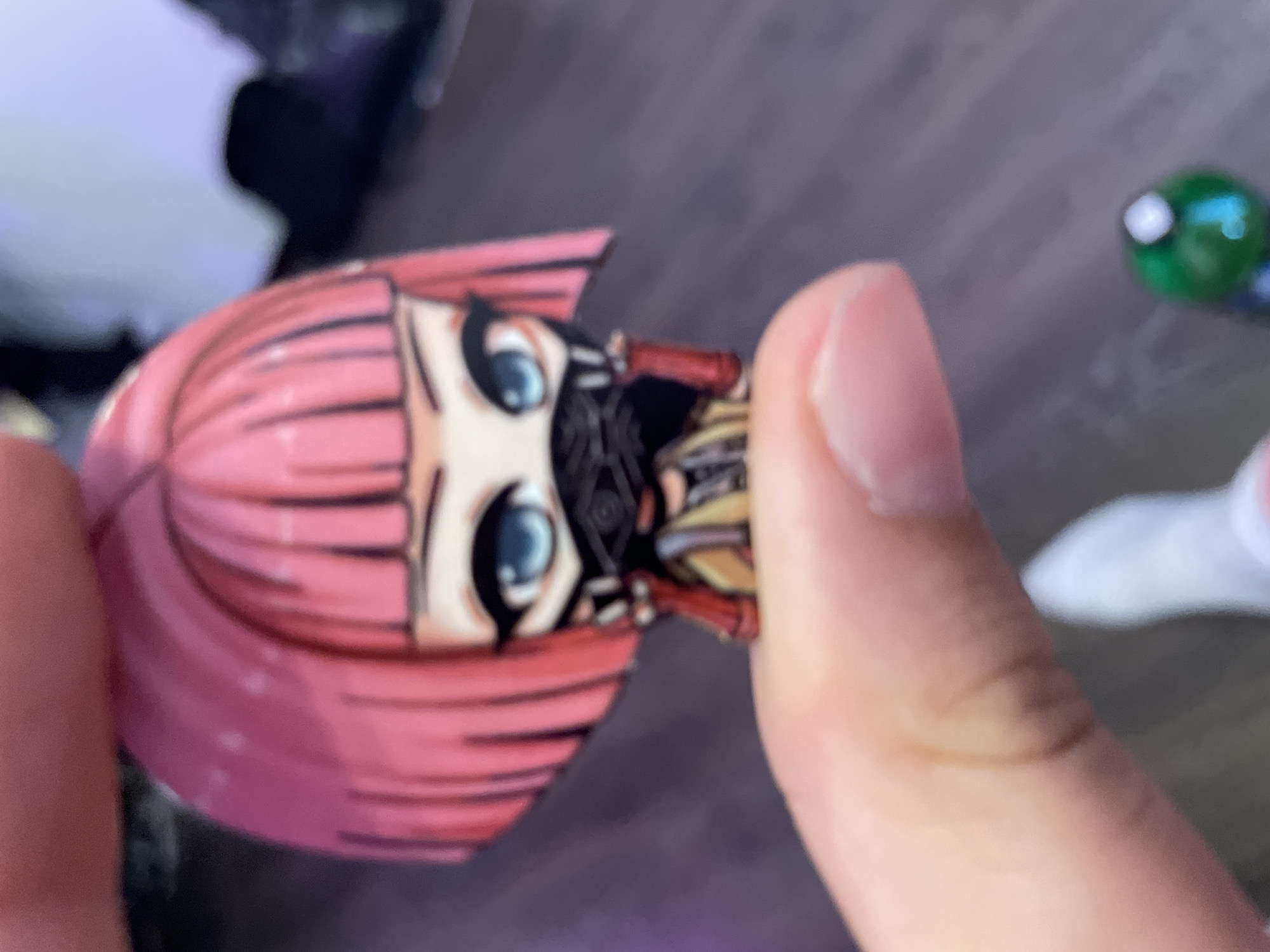 A close-up, slightly out-of-focus photograph shows a person's thumb and index finger holding a small, anime-style figurine of a character with pink hair, large blue eyes, and a black mask covering the lower half of her face. The figurine is depicted wearing a yellow and brown outfit with red accents. The background is blurred and appears to be indoors, with a dark, possibly wooden floor. There is a hint of a white shoe and a green object with a white top visible in the lower right corner of the image. The photograph was taken in the city of Sinyavino, Russia.