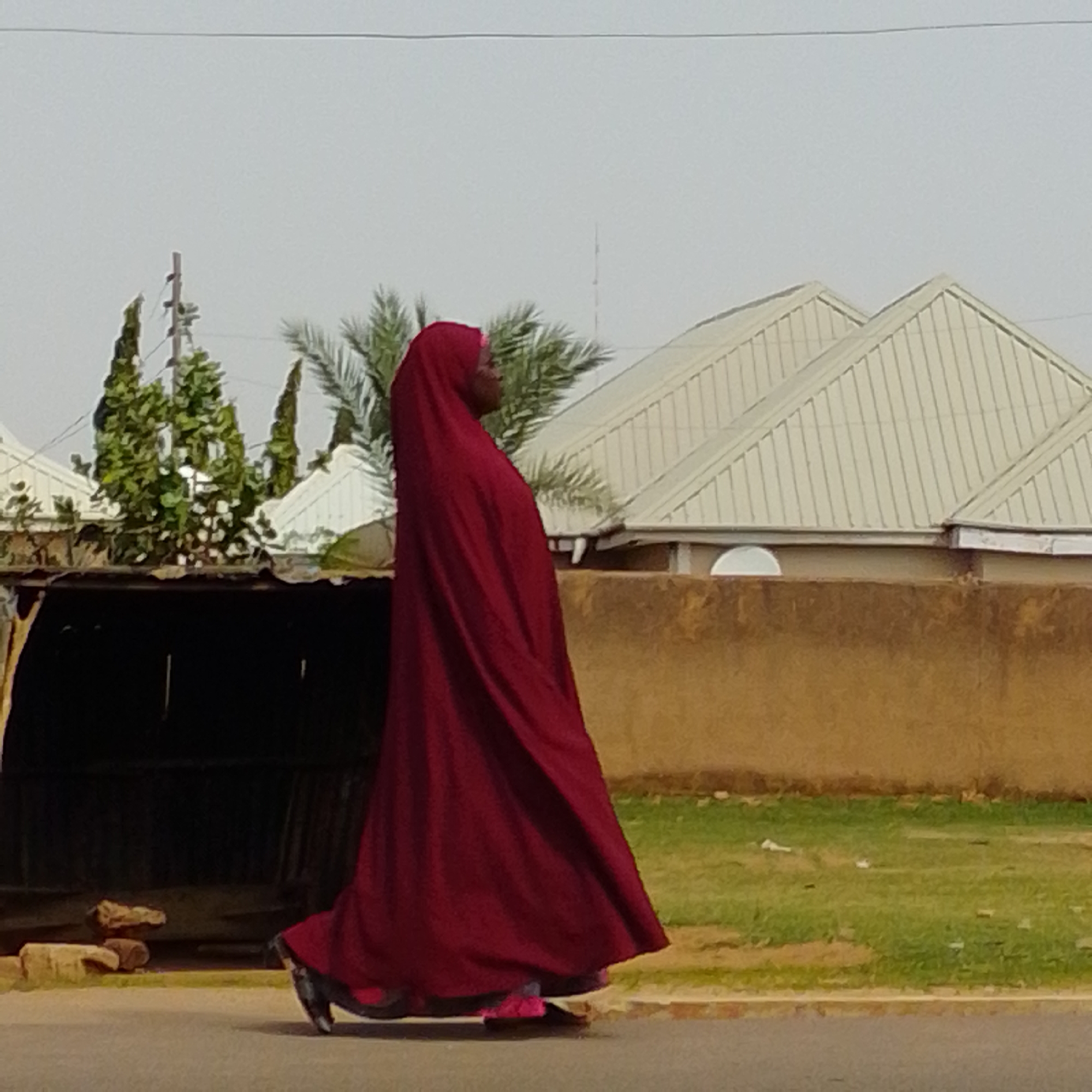 A full-shot, side-profile, medium-distance photograph depicts a person wearing a dark red Islamic hijab and dress walking on a paved road. The person is facing to the right. The hijab covers the person's head and hair, and the dress is long and flowing, reaching to the ground. The person's feet are visible, wearing dark shoes with pink accents. The background consists of a light brown, textured wall, a patch of green grass with some scattered white debris, and behind that, several white-roofed buildings with corrugated metal roofs. Trees, including palm trees, and utility poles are also visible in the background. The sky is overcast and light gray. The scene appears to be outdoors, possibly in a residential or commercial area.