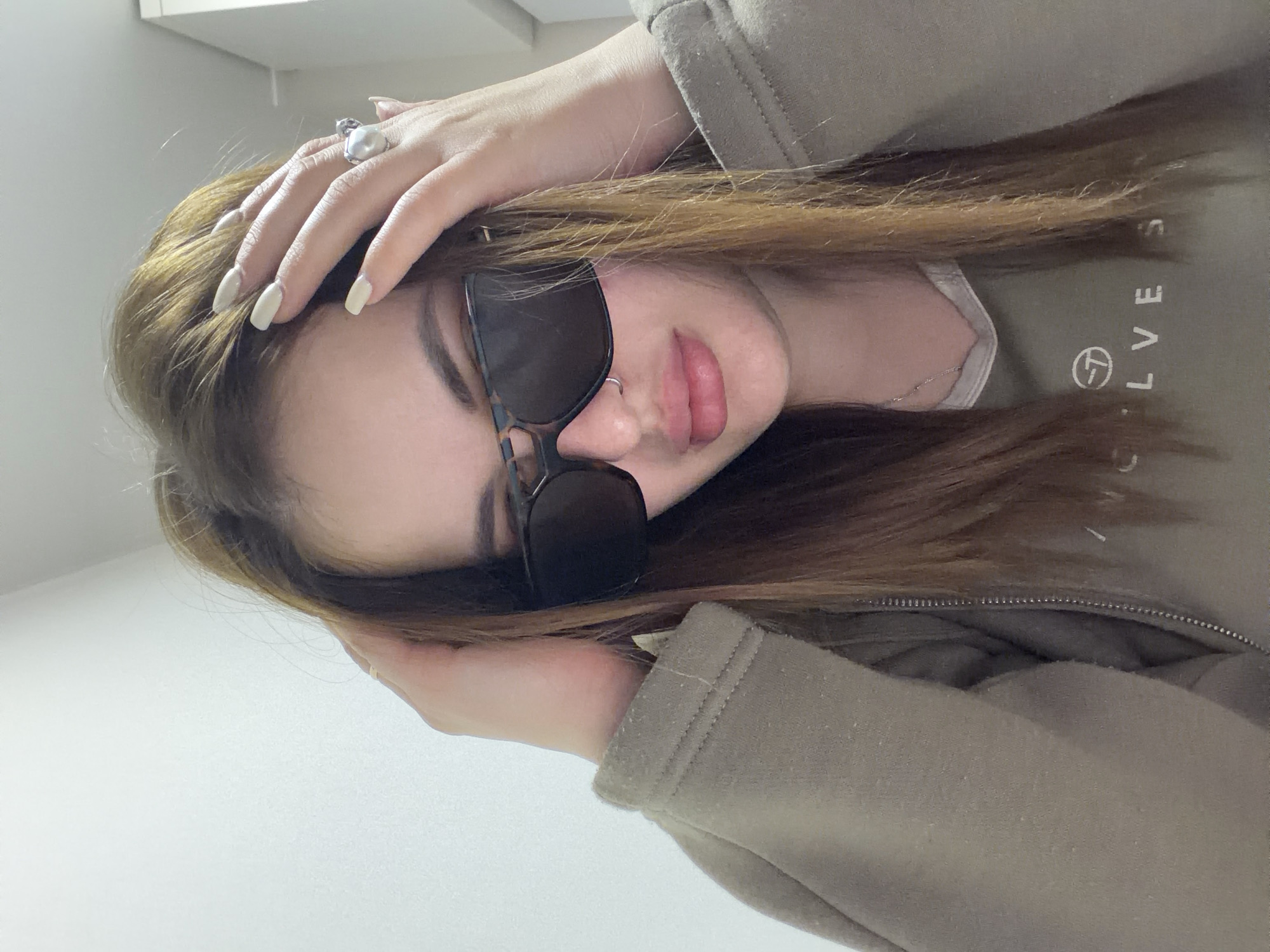 A young woman with light brown hair and wearing sunglasses poses indoors. She has a nose ring and is wearing a light brown zippered jacket with "C·LVES" printed on it. She is wearing a large ring on her hand. The background is plain white. This photo does not provide enough information to determine a specific location in Krasnodar, Russia.