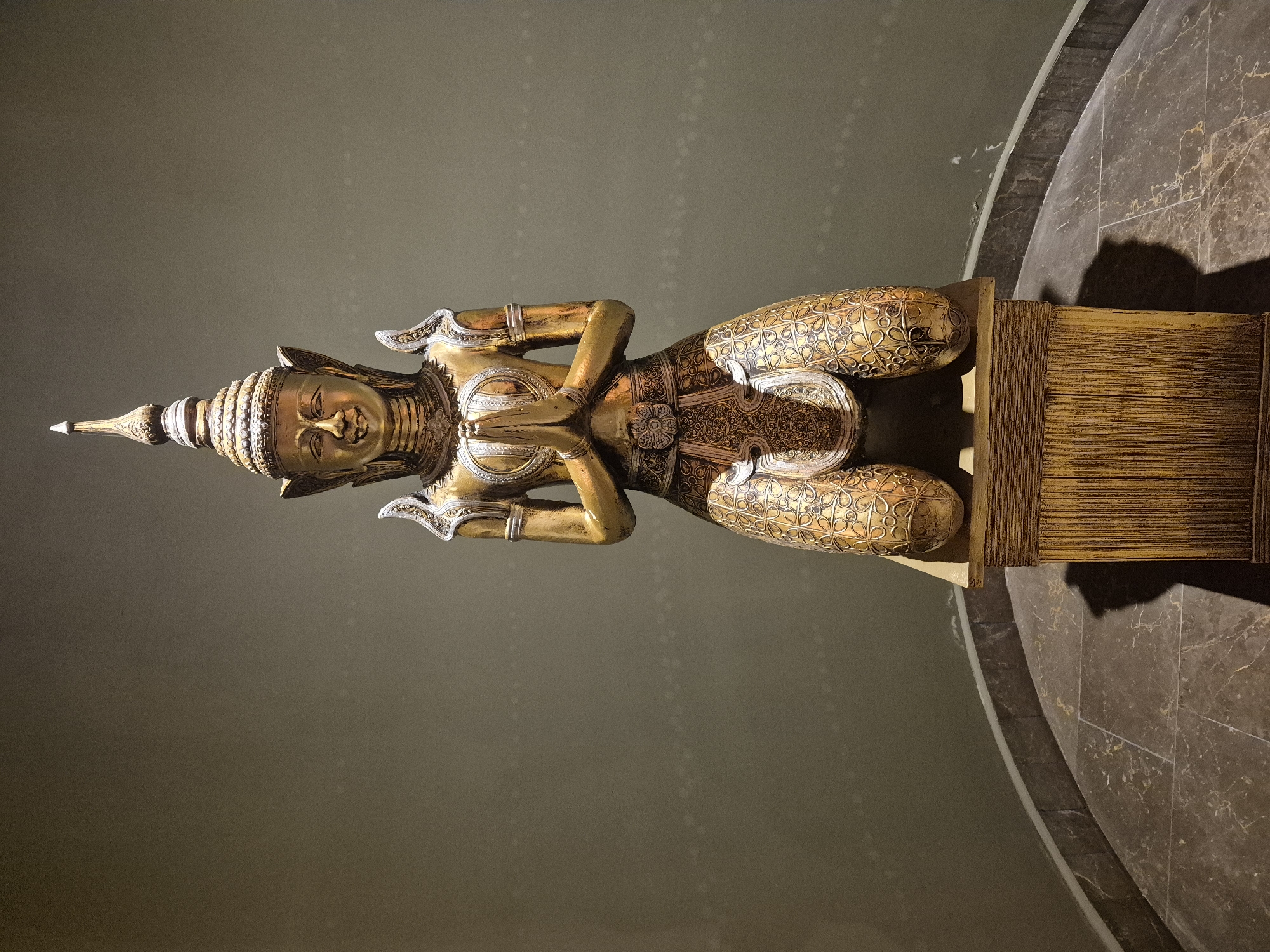 A full-body shot shows a statue of a Thai Buddha in a praying position on a pedestal in a museum in Alanya, Turkey. The Buddha statue appears to be made of gold with silver ornamentation and decoration. The Buddha is kneeling on the ground. Its head is covered in a Thai-style headdress. It has its hands folded together in a praying position. It is placed on a yellow pedestal that looks like a box. In the background is a green wall. To the right of the statue, the marble floor can be seen.