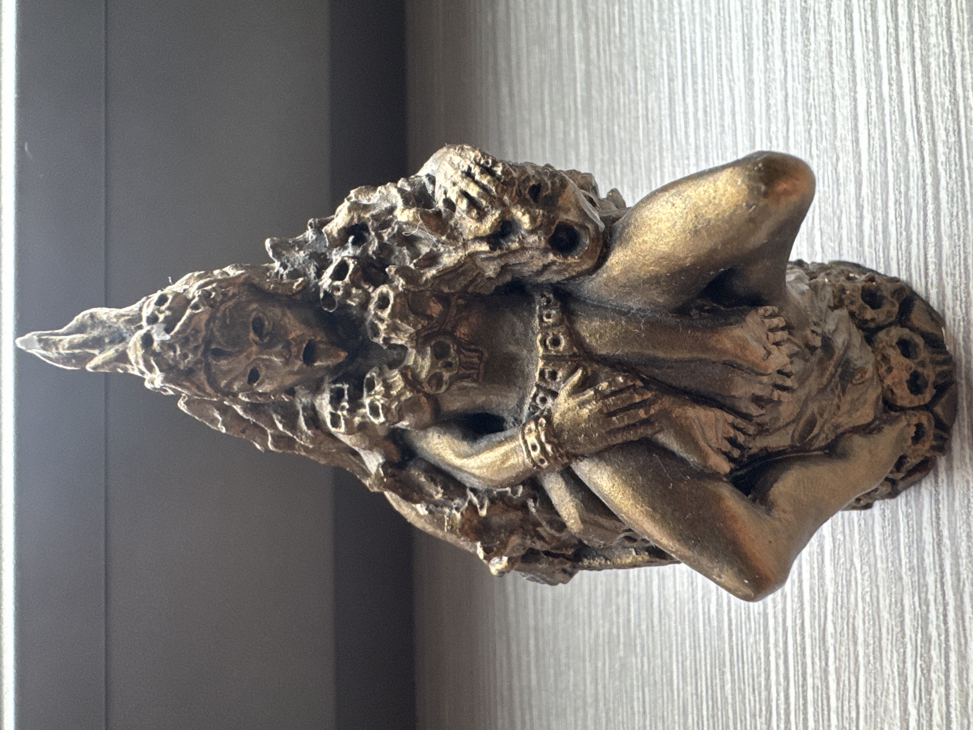This is a close-up, eye-level, front-facing photograph of a small, metallic statue. The statue is predominantly gold-colored and appears to depict a fearsome deity, possibly Kali, given its multiple arms, skull adornments, and aggressive posture. The figure has its mouth open and tongue extended, with a skull held in one of its hands. It is seated or crouched, with legs bent and one knee raised. The statue is placed on a flat, light-colored surface with a textured, wood-grain pattern. The background consists of a dark, out-of-focus area, possibly a wall or shelf, with a thin, bright horizontal line across the top. The lighting appears to be coming from the front and slightly from above, casting subtle shadows. The image was taken indoors, and the overall impression is of a still-life object display. There is no indication of the city of Konakovo, Russia, in the image itself or its context.