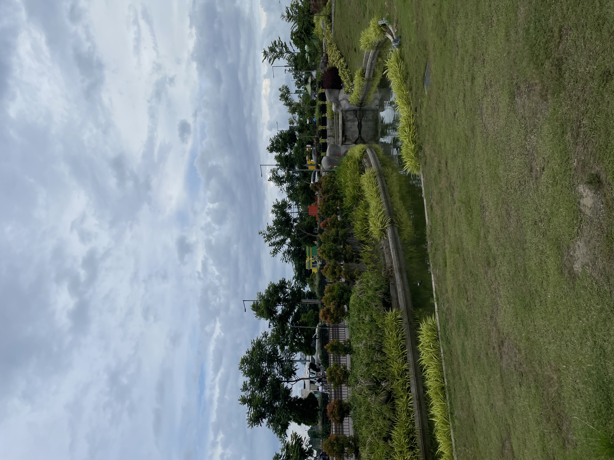 The image depicts an outdoor scene in a park or landscaped area, likely in Davao City, Philippines, given the context. The sky is overcast with a gray, cloudy expanse dominating the upper portion of the frame. In the middle ground, a man-made water feature, possibly a canal or pond, is visible. It is lined with lush green vegetation, including bright yellow-green ornamental plants. Beyond the water feature, there is a paved area with vehicles and people, suggesting a public space like a road or parking lot. Several trees with dense green foliage are scattered throughout the middle ground and background, some with streetlights nearby. The foreground is a grassy slope with patches of darker soil visible, indicating a well-maintained but naturalistic park setting. The overall impression is one of an urban green space on a cloudy day.