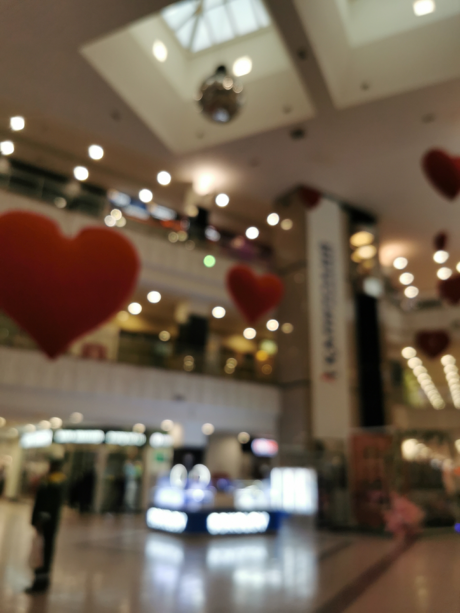 The image shows the interior of a shopping mall. There are red heart decorations hanging from the ceiling. In the foreground, there is a person in a dark coat walking. There are also display cases or kiosks with bright lighting. A skylight and a disco ball are visible on the ceiling. A tall column with unreadable text or graphics is in the middle ground. Based on the presence of a disco ball and the upscale design of the interior architecture, the image might depict the GUM department store on Red Square in Moscow, Russia, known for its festive decorations. The image is notably blurred, which makes discerning details challenging.