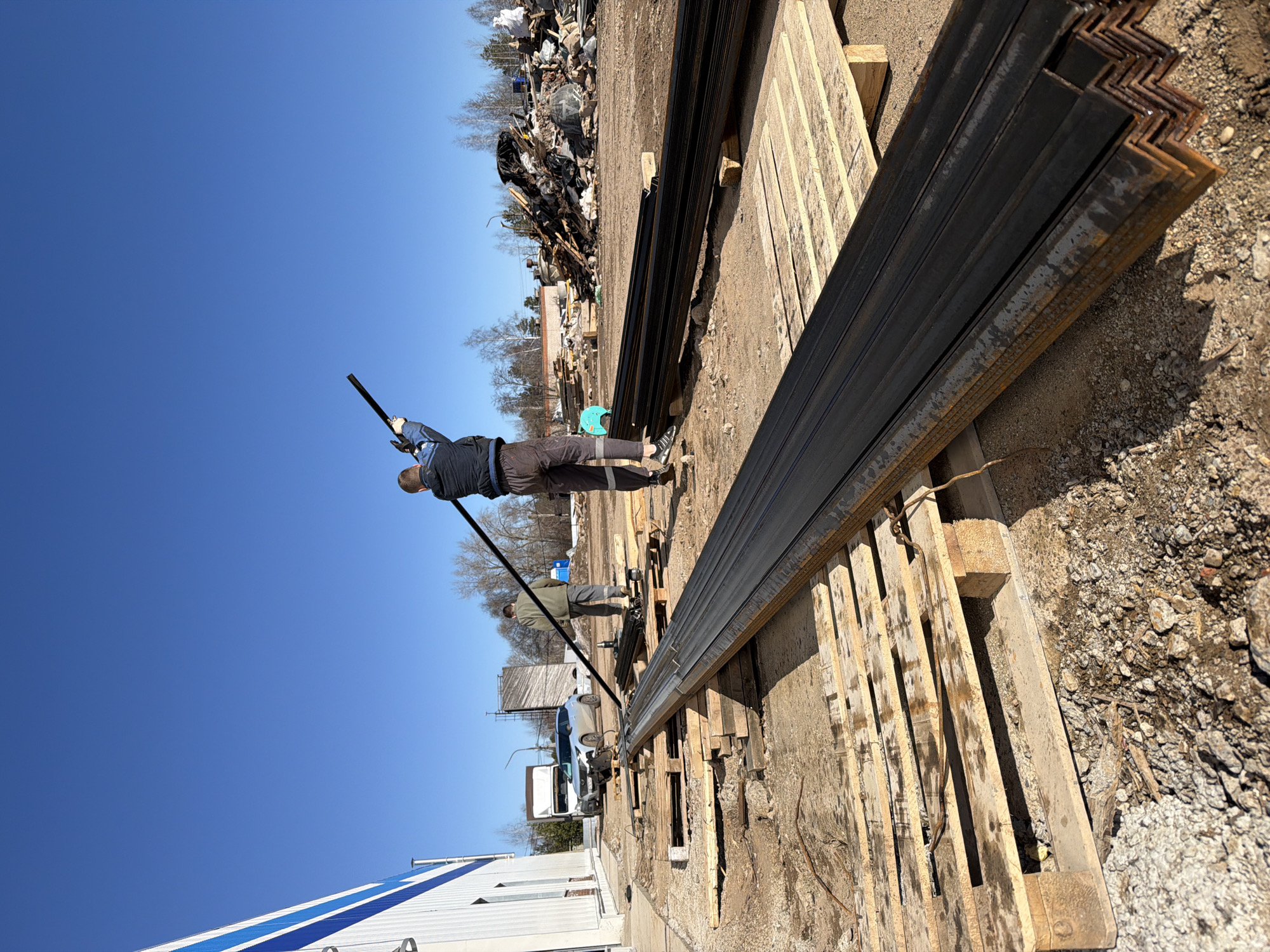 Location: Chaikovsky, Russia.
Scene: A salvage yard/industrial area. A clear, blue sky is overhead. Two men are present. One man, wearing a blue jacket and dark pants, stands near a stack of L-shaped metal bars, holding a long black pole. Another man, wearing a green jacket and dark pants, stands further away. The metal bars are stacked on wooden pallets. In the background, there is a pile of discarded materials (trash), a white truck, a silver car, and a partially visible building. The ground is a mix of dirt and gravel.