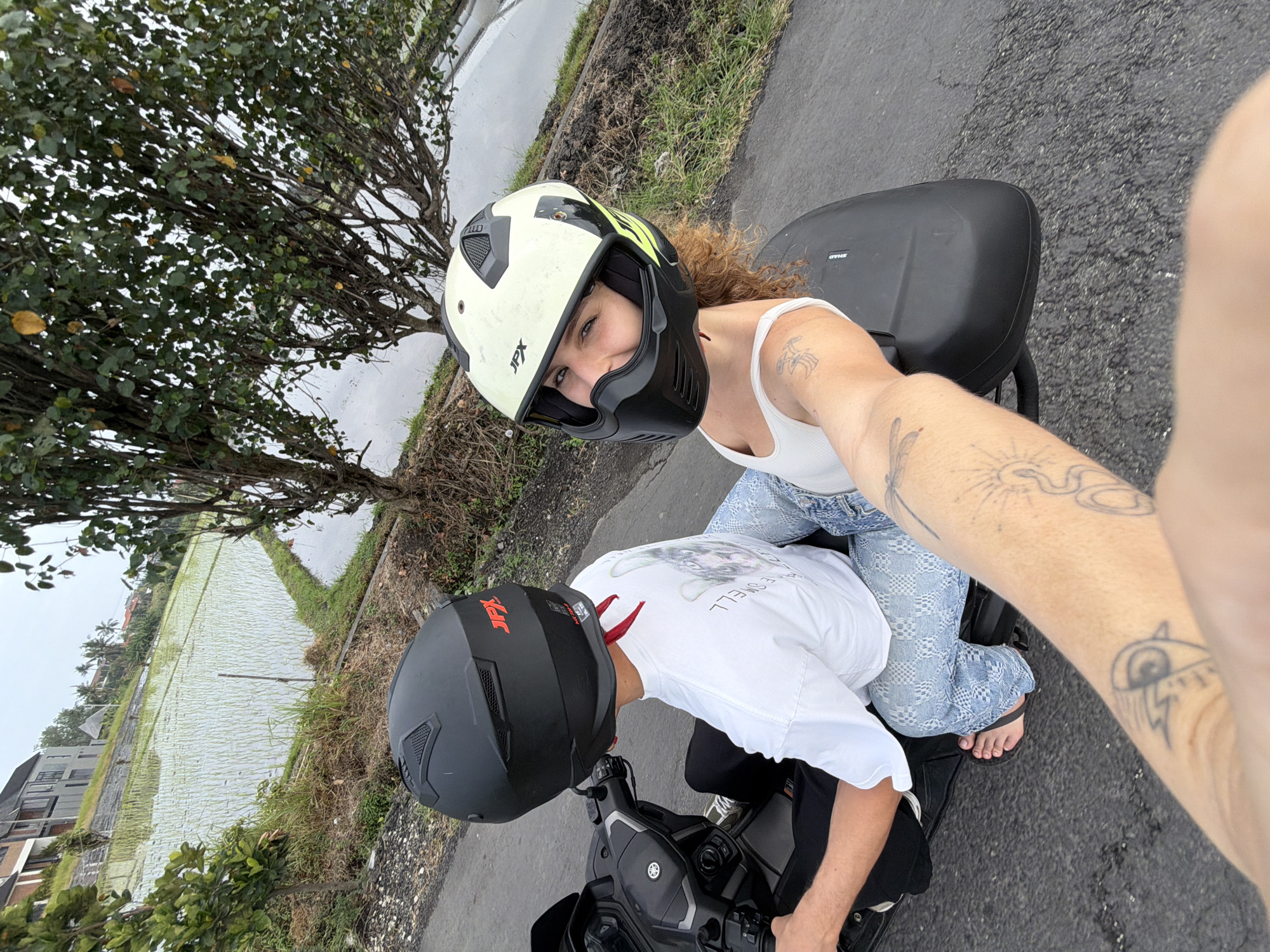 In Mengwi, Indonesia, two individuals are captured on a motorcycle. The person in the front, wearing a black helmet, is operating the vehicle and is dressed in a white t-shirt and blue jeans. The person in the back, with curly hair, wears a white and black helmet and a white tank top. Their arm extends towards the camera, displaying tattoos on their forearm. They are situated on a paved road next to a lush green rice paddy. In the background, modern buildings and trees are visible, suggesting a suburban or rural setting. The sky appears overcast, indicating a possibly humid climate. The scene is photographed from a perspective that suggests it was taken while the motorcycle was in motion.