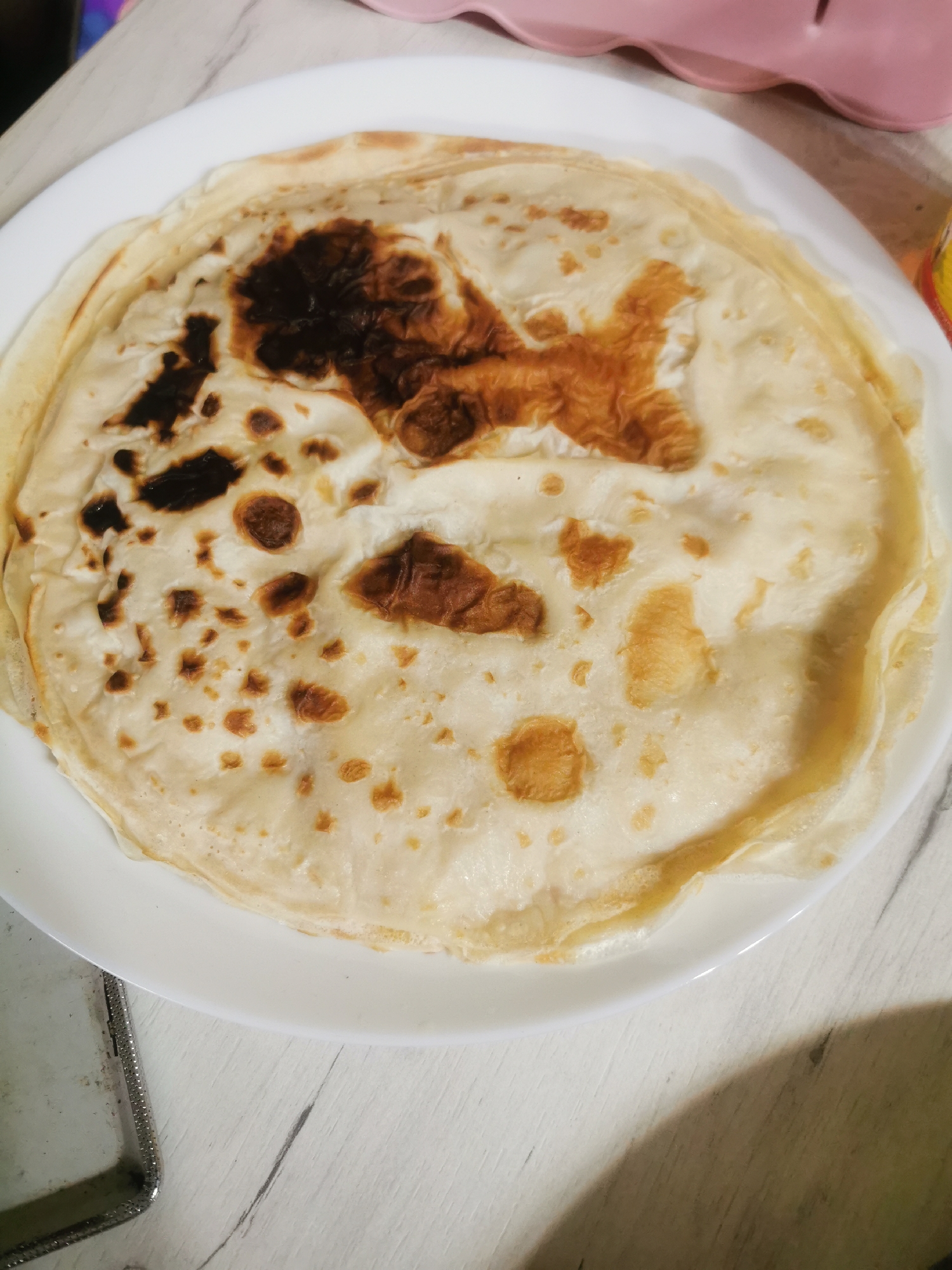On a white plate, there is a stack of what appears to be crepes or thin pancakes. The pancakes have a light, slightly uneven color with some darker brown patches, indicating they were cooked on a hot surface. They are stacked in a circular shape on the plate. The plate sits on a light-colored, possibly wooden, surface. To the left of the plate, there is a glimpse of a metallic object, possibly part of a device. In the background, upper-right corner, there is a pink-colored container, out of focus. The photograph is taken from an overhead perspective. There is nothing to suggest this is from the city of Stara Zagora, Bulgaria.