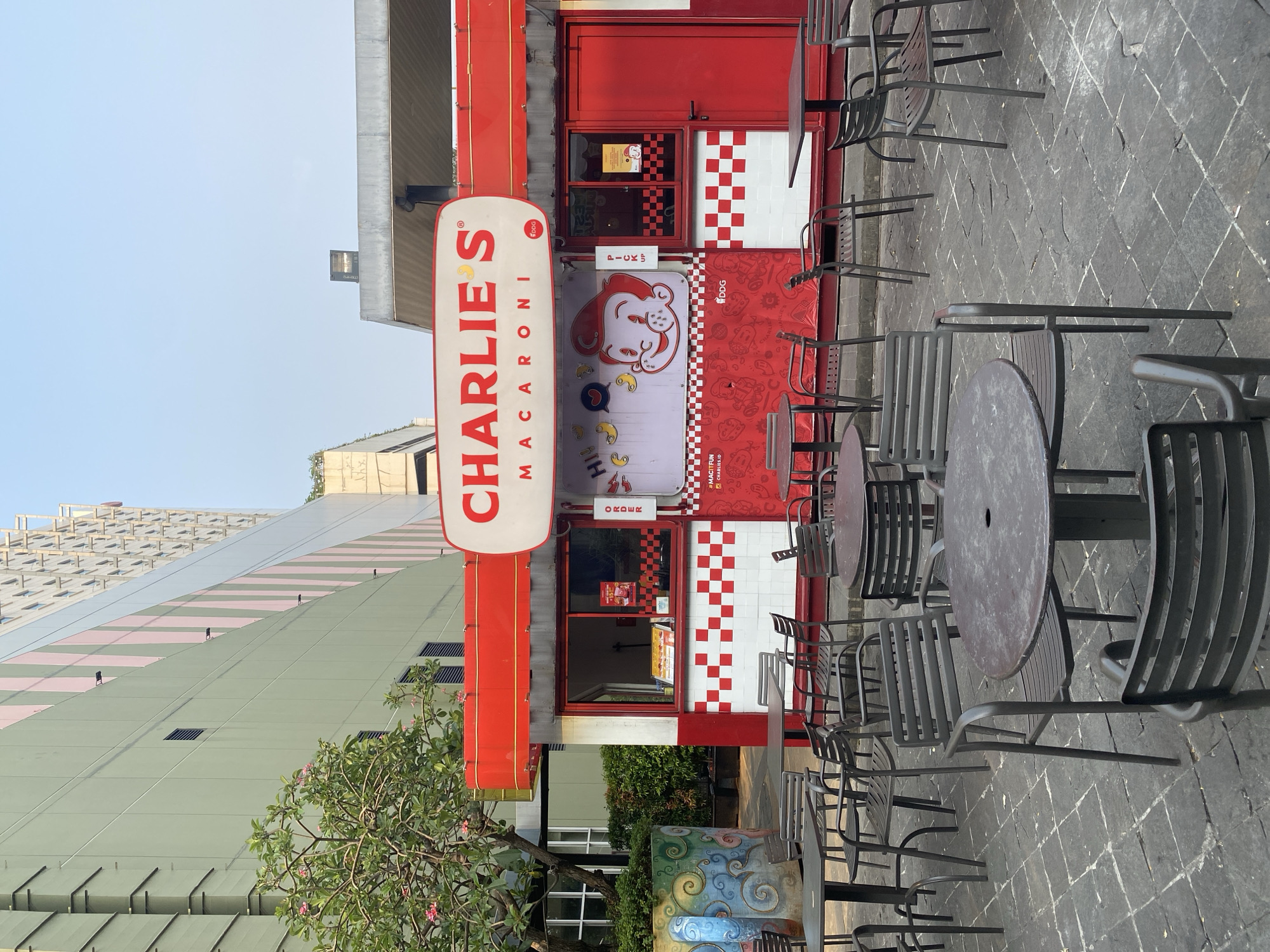 A red and white food stall named "CHARLIE'S MACARONI" is depicted in an outdoor seating area in North Jakarta, Indonesia. The stall has a bright red facade with white checkerboard accents and a prominent sign. Several metal tables and chairs are arranged on a stone-tiled ground in front of the stall. Behind the stall, a modern building with green and pink vertical panels is visible, suggesting an urban setting. The sky above is clear and blue. There are no people visible in the image.
