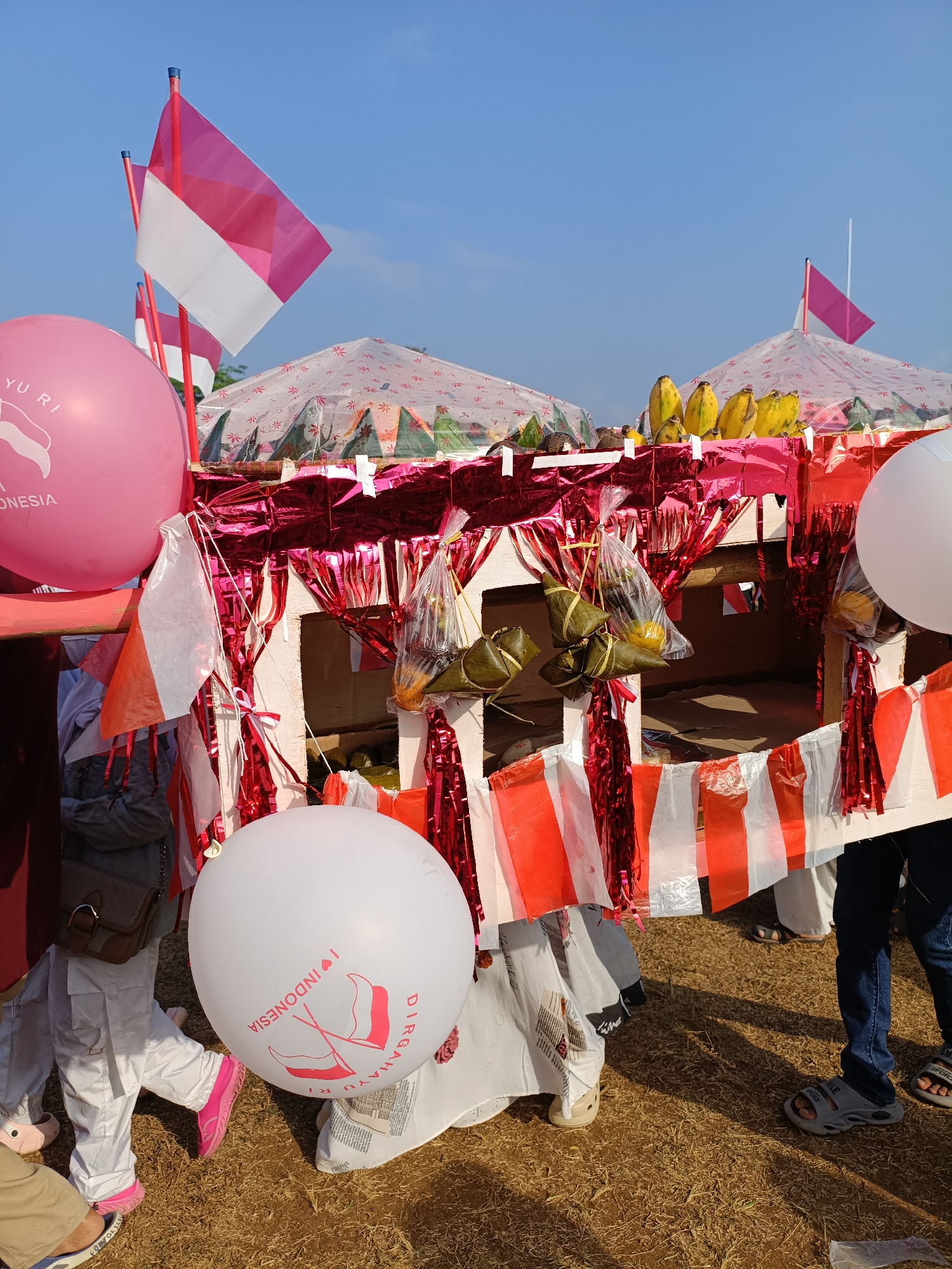 The image depicts an outdoor scene in Cibodas, Indonesia, likely during a celebratory event. Several Indonesian flags, which are red and white, are prominently displayed, attached to poles and as small pennants. There are large pink and white balloons, some with text and imagery related to Indonesia, indicating a patriotic or nationalistic theme. In the background, tents with patterned coverings are visible, suggesting a festival or market.
The foreground features a decorated stall or structure adorned with red and white streamers. Several bunches of bananas are displayed on top of this structure, and some food items, possibly wrapped in banana leaves and plastic bags, are hanging from it. The ground is covered with dry grass or straw, and several people are partially visible, their lower bodies and legs showing. They are wearing casual clothing and sandals. The overall atmosphere appears festive and public. The presence of Indonesian flags and thematic balloons strongly suggests a celebration of Indonesian independence or a related national holiday.