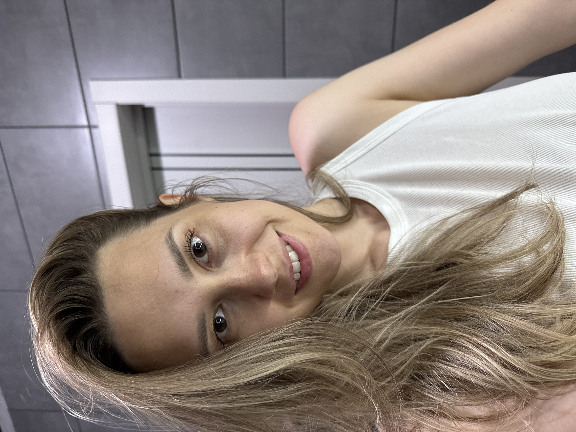 A woman with light brown hair and brown eyes smiles at the camera. She is wearing a white tank top. The background includes a gray tile wall and a white door frame. The image appears to have been taken indoors, possibly in a bathroom. There is no information available to confirm the location in the city of Chaikovsky, Russia.