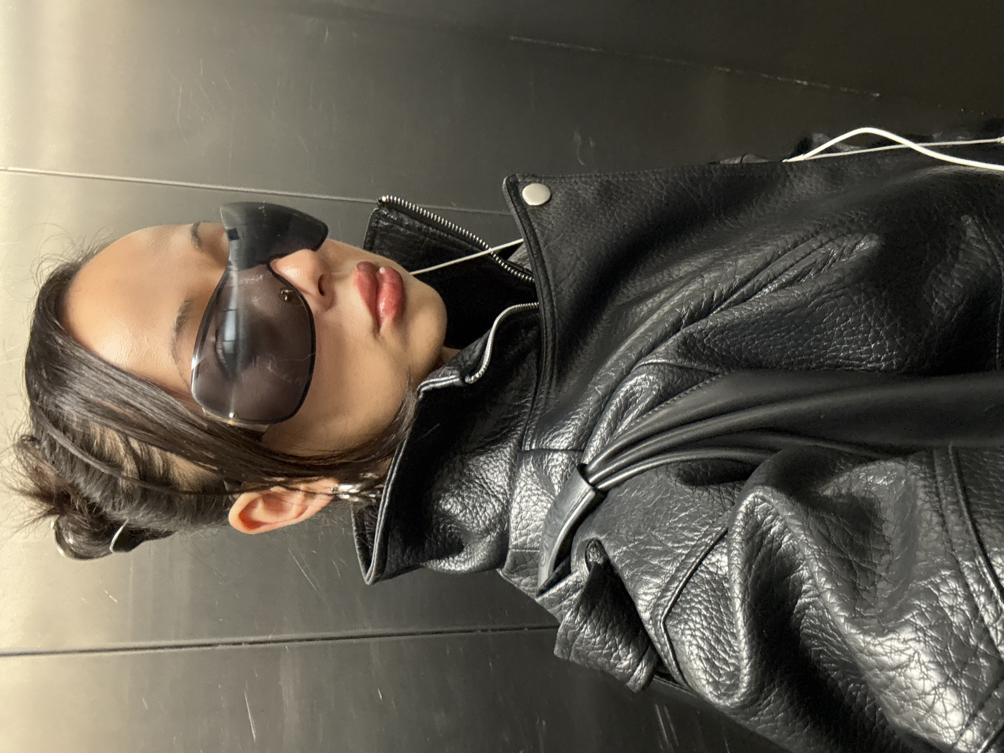 A close-up portrait shot, rotated 90 degrees counter-clockwise, features a person, likely female, wearing a stylish black leather motorcycle jacket. The jacket has a pebbled texture, visible creases, a zippered front, and silver snap buttons on the lapels. The person has dark hair pulled back, revealing an ear with a white earbud in place, connected by a visible white wire draped over the jacket. Dark, possibly futuristic-style sunglasses with a thick nose bridge are worn, obscuring the eyes. The person's lips are full and tinted reddish. They are posing with their head tilted slightly back and looking upwards. The background is a plain, dark, possibly metallic or tiled wall, showing faint horizontal lines and minor scuffs, suggesting an urban indoor environment such as a hallway, elevator, or subway station. The overall mood is cool and confident. No text is visible in the image. The setting is consistent with an urban context like London, United Kingdom.