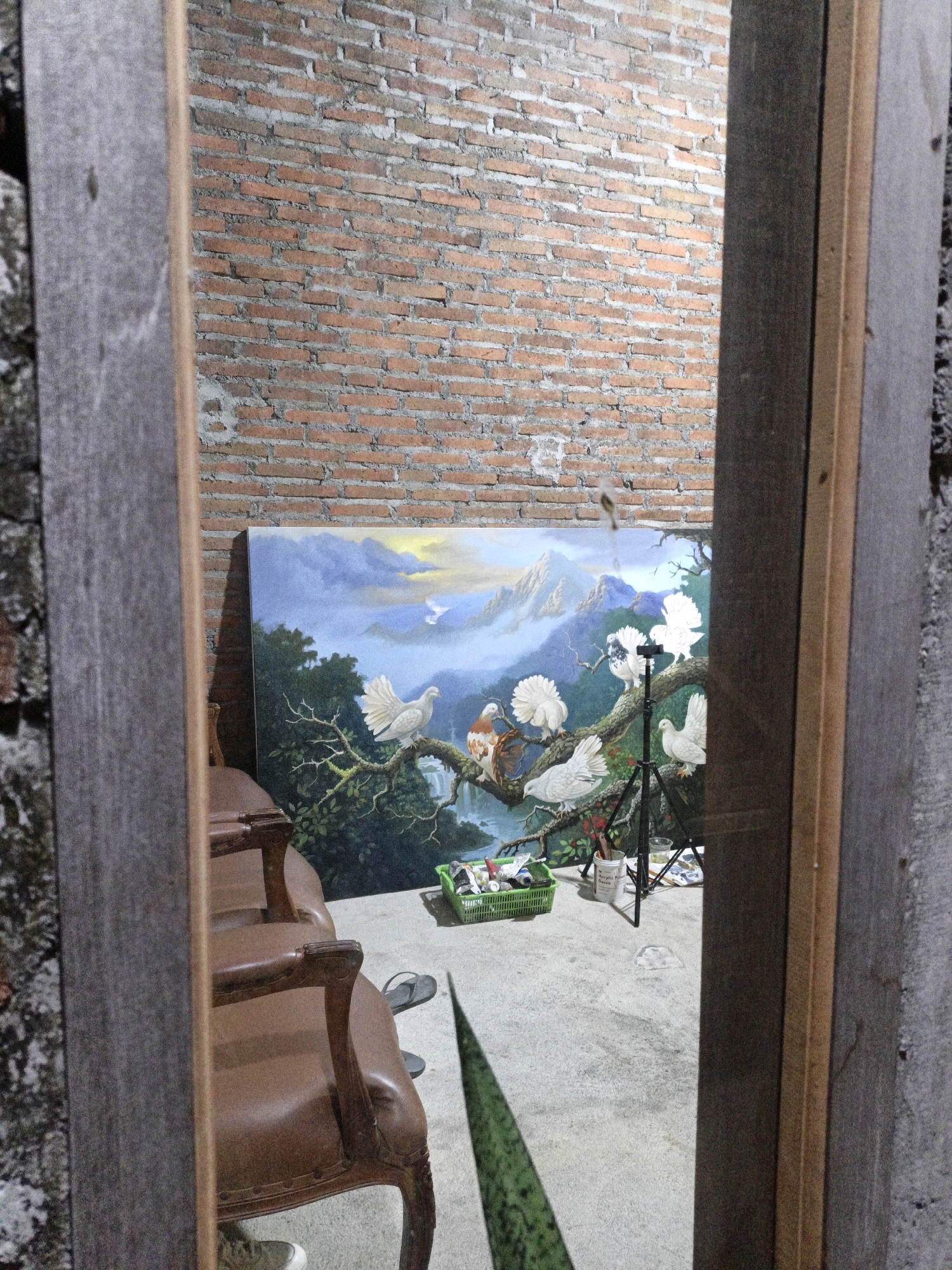 The image shows a painting of birds on a branch against a mountainous backdrop. The painting is reflected in a mirror, which is framed by wooden elements. In the foreground, to the left, is a brown leather armchair. To the right, a portion of the mirror frame is visible. In the background, a brick wall is evident. A green basket filled with art supplies, including tubes of paint, is placed on the floor in front of the painting. The scene appears to be indoors, possibly in a studio or workshop, with a concrete floor. No people are present in the image. The location in Cikal, Indonesia, is not discernible from the visual information provided in the image.