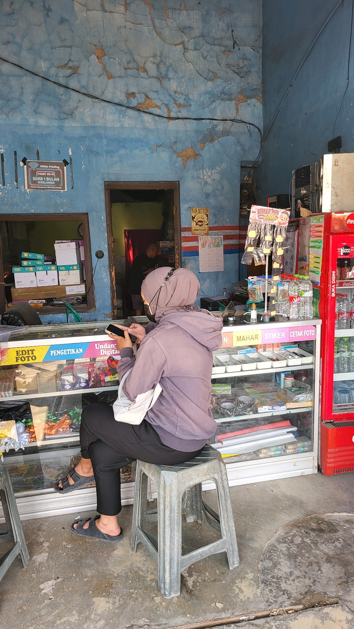 In the city of Lebaksiu, Indonesia, a woman wearing a hijab and a purple hoodie sits on a gray plastic stool in front of a storefront. She is holding a smartphone in her hands and appears to be looking at it. The storefront is stocked with various items, including what appears to be stationery, drinks in a red refrigerator, and other miscellaneous goods. The walls of the building are painted blue and show signs of age and wear, with peeling paint and cracks. A doorway in the back of the storefront leads to an interior area where another person is visible. The ground in front of the store is concrete with some debris and water stains.