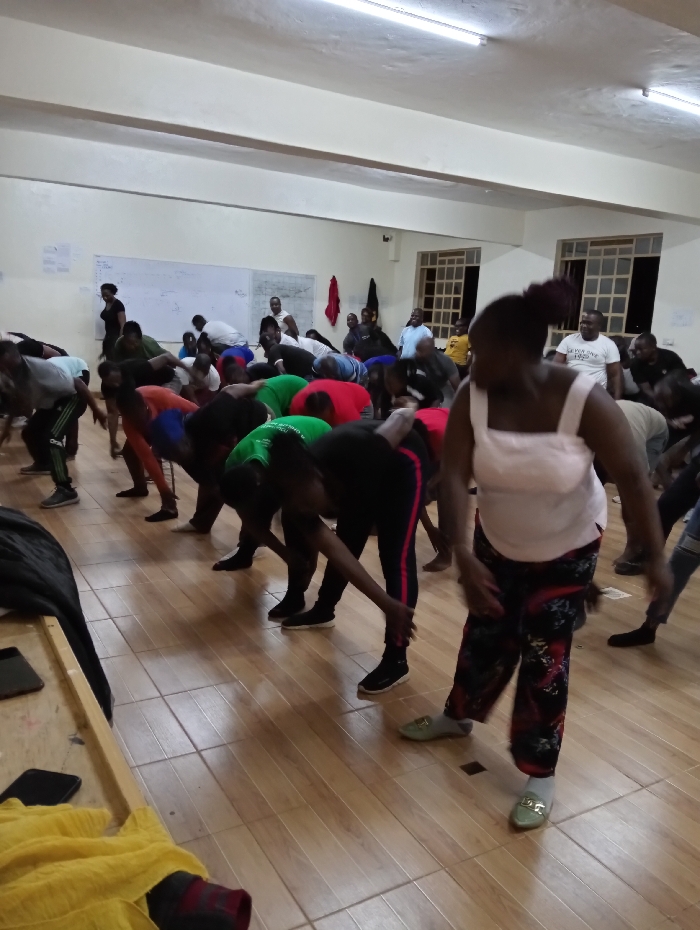 An indoor scene, likely a community hall or exercise studio in Riara Ridge, Kenya, depicts a large group of adults engaged in a group physical activity. The room features light-colored walls, a light wooden tiled floor, and ceiling-mounted fluorescent lights. On the left wall, a whiteboard with some faint, illegible markings is visible, while two windows with grilles on the right side appear dark outside, suggesting the event is taking place in the evening or at night.
Most participants are actively performing a movement, bent forward at the waist with their hands reaching towards their feet or the floor, indicative of stretching, exercise, or a dance routine. They are dressed in casual and comfortable attire, including t-shirts, track pants, and a mix of athletic shoes and casual footwear. In the immediate foreground on the right, a woman wears a white tank top, patterned trousers, and light green flat shoes. Another individual in the mid-ground left is dressed in a black top and black and red striped pants, also in a bent posture.
The atmosphere suggests focused participation, with individuals concentrating on the collective movements. A wooden table or bench draped with yellow fabric is partially visible in the foreground on the left. No clear, legible text is visible within the image.