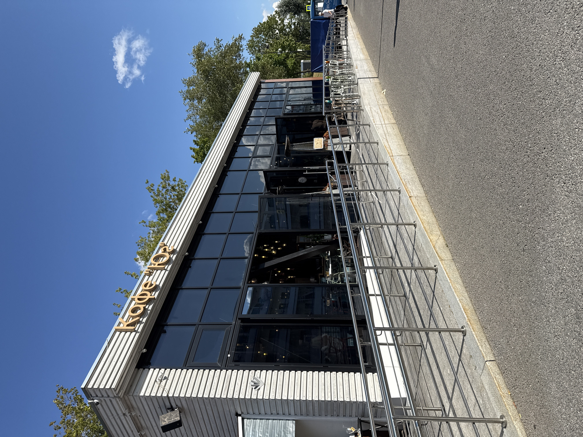 This is a medium-angle exterior shot of a modern cafe, labeled "Кафе "102"", in Khimki, Russia. The cafe has a large, glass-fronted facade with multiple doors, suggesting an open and inviting atmosphere. The building's exterior is clad in horizontal white siding. A ramp with a stainless steel railing leads up to the entrance, indicating accessibility. Inside, cafe tables and chairs are visible, along with decorative lighting. The environment is bright and sunny, with a clear blue sky and trees in the background. A few people are present, some standing near the entrance and others seated at outdoor tables. The overall scene depicts a casual, urban dining establishment on a pleasant day.
