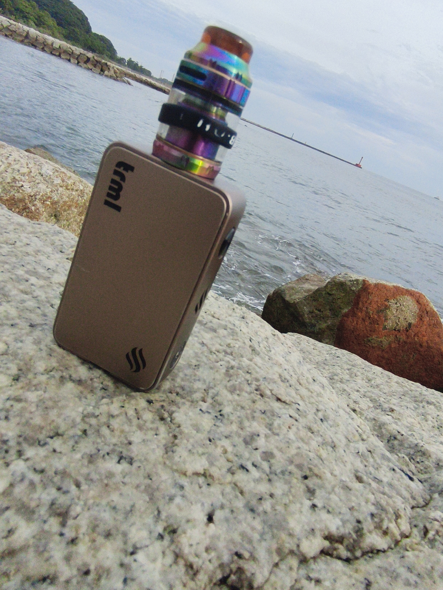 An electronic cigarette device rests on speckled granite rocks near a body of water. The device, a bronzed box mod with a rainbow-colored atomizer, is positioned at an angle. In the background, the sea stretches out, meeting a pale, cloudy sky. A man-made structure, possibly a breakwater, extends into the water, and a distant red buoy is visible. Lush green foliage crowns the land beyond the breakwater. The scene appears to be a coastal area, possibly in Lhokseumawe, Indonesia, given the typical geological formations and coastal environment.
