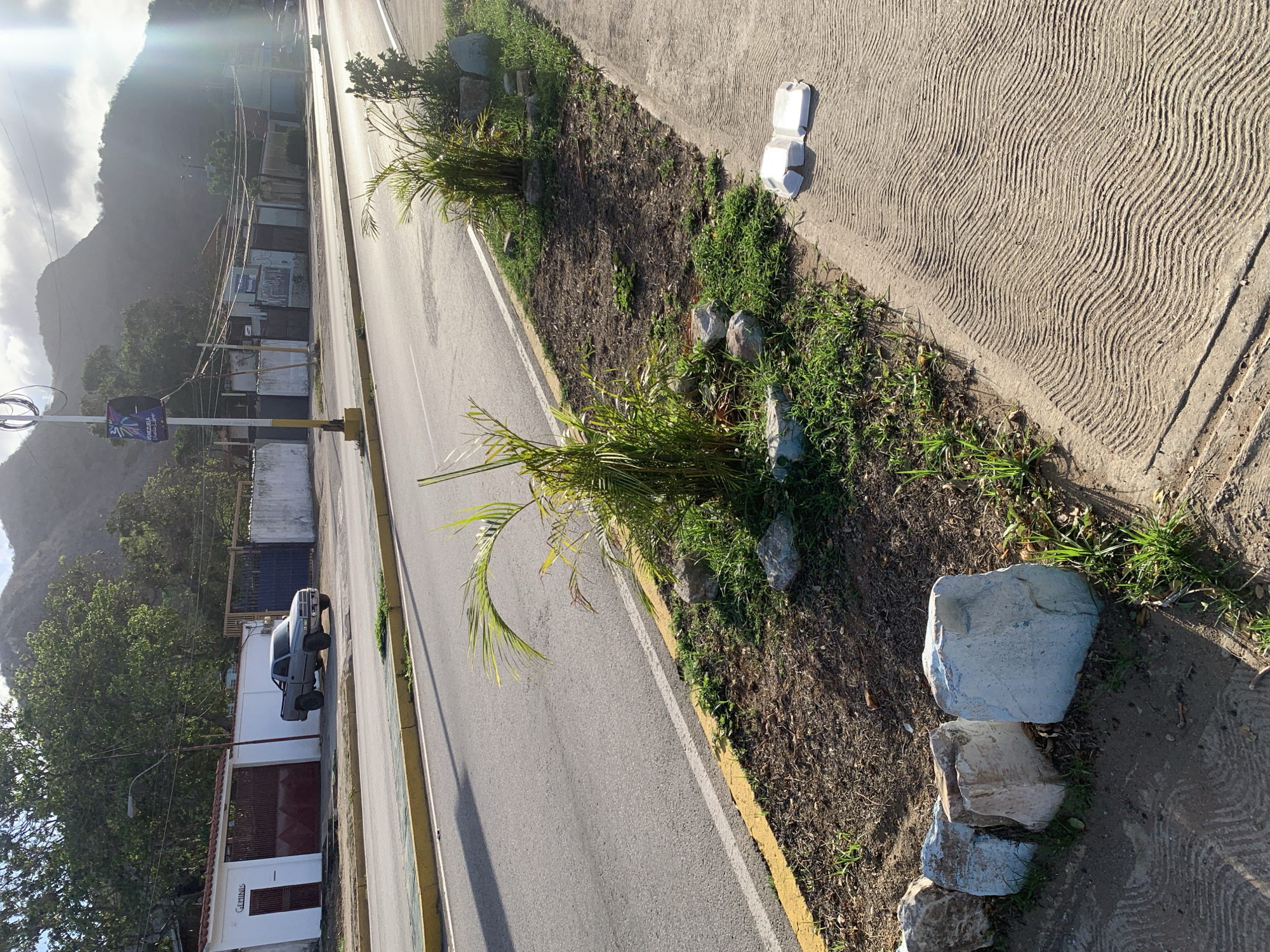 The image shows a roadside scene in El Valle del Espíritu Santo, Venezuela. On the left, a gray pickup truck is parked in front of a white building with a large, dark red metal gate. The building has a smaller, dark red door on the left side, and text above it that reads "GEMINIS". A lamppost stands in front of the building. Beyond the building, more structures and fences are visible. The road in the foreground is asphalt with a yellow line along the edge. A strip of dry earth and sparse grass runs along the side of the road, bordered by large rocks and some young palm trees. On the right side of the road, a concrete sidewalk with a wavy pattern is visible, with two white styrofoam containers placed on it. In the background, mountains rise under a hazy sky. The sun is low, casting long shadows and creating lens flare.