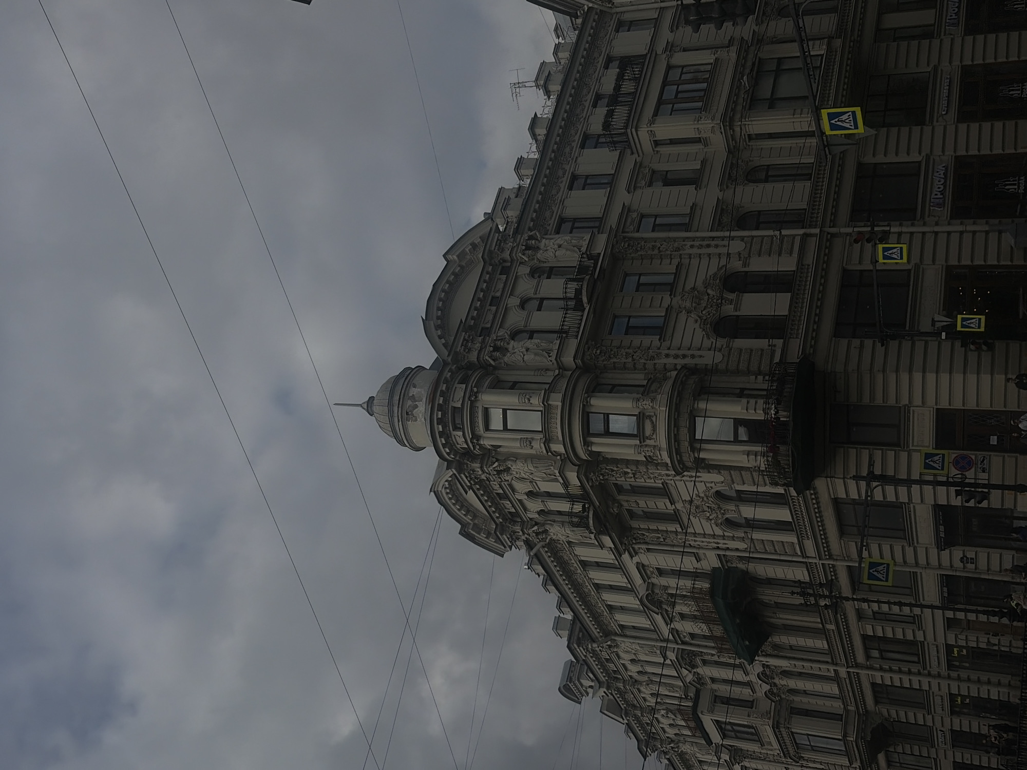 The image depicts a street view of an ornate, multi-story building in Saint Petersburg, Russia, under a heavily overcast sky. The architecture is detailed and classical, with numerous windows, balconies, and decorative elements. The building is the corner of a street, with another building partially visible on the right. Several power lines crisscross the sky above the buildings.
In the foreground, there are streetlights, pedestrian crossing signs with yellow backgrounds and pedestrian icons, and traffic lights. The bottom of the frame shows the sidewalk and the lower floors of the buildings, which appear to be shops or businesses, with some text visible on their facades, including "ГЛОБАЛ" and "ПОЧТА". A few people are faintly visible on the sidewalk. The overall atmosphere is somber due to the dark clouds and the muted color palette.