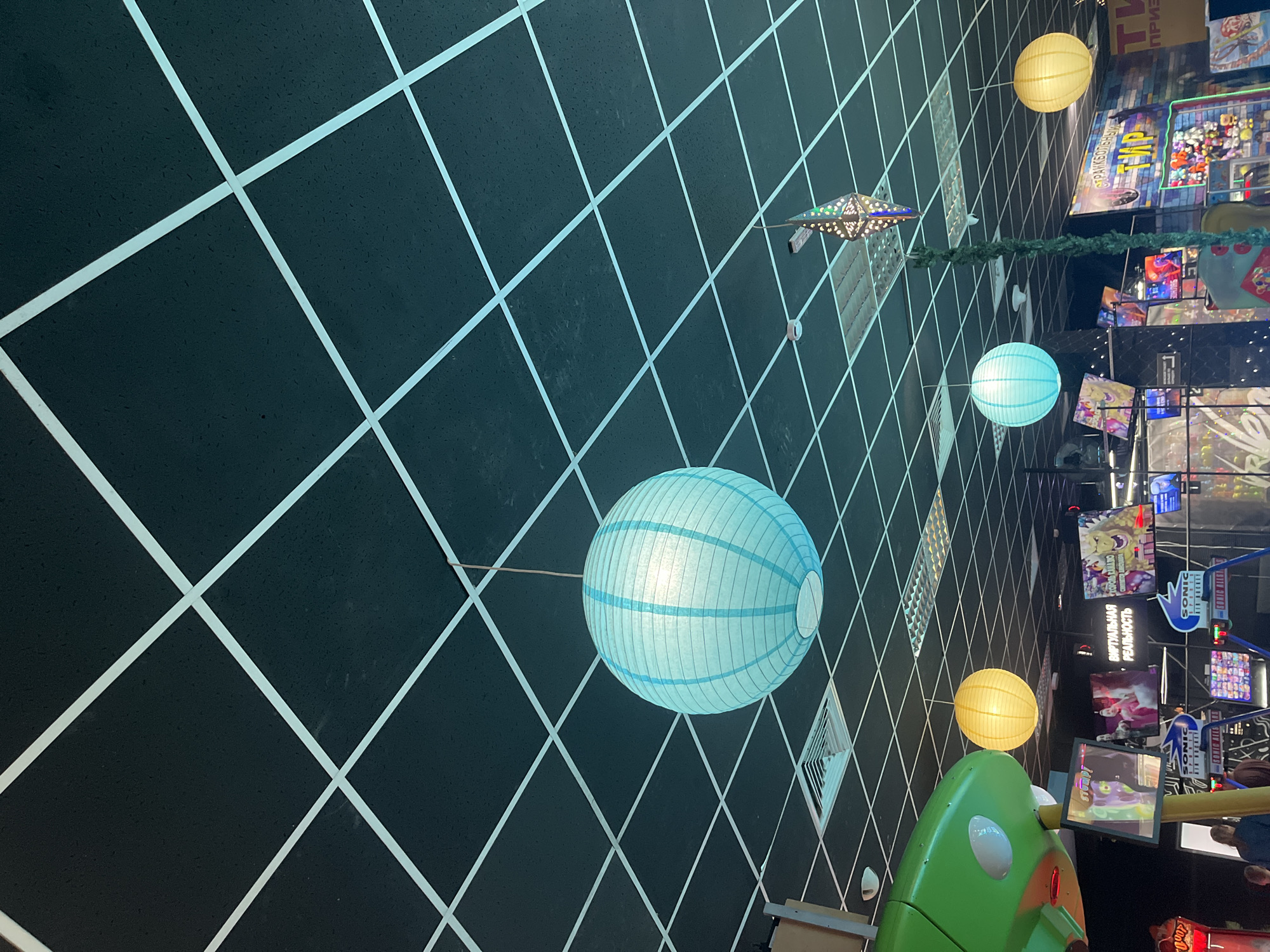 The image displays an indoor entertainment venue, likely an arcade or family center, characterized by a distinctive ceiling or wall design. A dark, possibly black or very dark grey, surface is adorned with a bright white grid pattern, creating a strong sense of perspective and depth, reminiscent of a digital or futuristic environment.
Several decorative elements are suspended within this space. A prominent large, light blue paper lantern with horizontal stripes hangs in the mid-ground. Further back, another smaller light blue lantern and two yellow/orange spherical paper lanterns are visible. A unique star-shaped metal lantern, embellished with colorful lights, adds an exotic touch, while a green artificial garland or vine is strung horizontally across the scene.
In the background, numerous brightly lit arcade machines and display screens showcase colorful game graphics. Visible text on signs and screens confirms the setting as an entertainment hub in Russia. Text in Cyrillic includes "ВИРТУАЛЬНАЯ РЕАЛЬНОСТЬ" (VIRTUAL REALITY) on a black sign, and "СТРАЙКБОЛЬНЫЙ ТИР" (Airsoft Shooting Gallery) on another. The English word "SONIC" also appears on some game cabinets.
A large, rounded green structure, possibly part of an arcade game or play equipment, is partially visible in the foreground (bottom left of the original image). The scene is devoid of people, but the vibrant lighting and colorful displays suggest a lively and engaging atmosphere typical of a recreation area. The overall impression is one of fun and playful excitement.