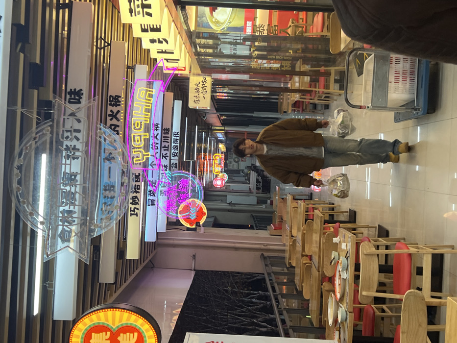 The image captures an indoor commercial walkway or food street in Jiangjin, China, under bright artificial illumination. A young East Asian male, appearing to be in his late teens or early twenties, walks forward, slightly right of center. He wears a brown bomber jacket over a light cable-knit sweater, light blue jeans, and tan shoes. He holds a translucent plastic takeout bag in each hand. His mouth is slightly ajar, and his gaze is directed ahead.
To the left, a row of light wooden tables with red-seated chairs lines a dining area, separated from an exterior space by a metal railing. Several tables in the foreground display remnants of meals, including bowls and plates. Beyond the railing, bare tree branches are visible against a dark, indistinct background.
The ceiling above is adorned with numerous illuminated signs in Chinese characters, some featuring neon elements, including a prominent pink/purple neon sign spelling "YUQIA." Other signs are round or arrow-shaped. The right side of the walkway features storefronts, some with glass facades. A utility cart with a blue base and white plastic crates is partially visible in the lower right foreground, next to a person's obscured shoulder and arm. The floor is light-colored and reflective.