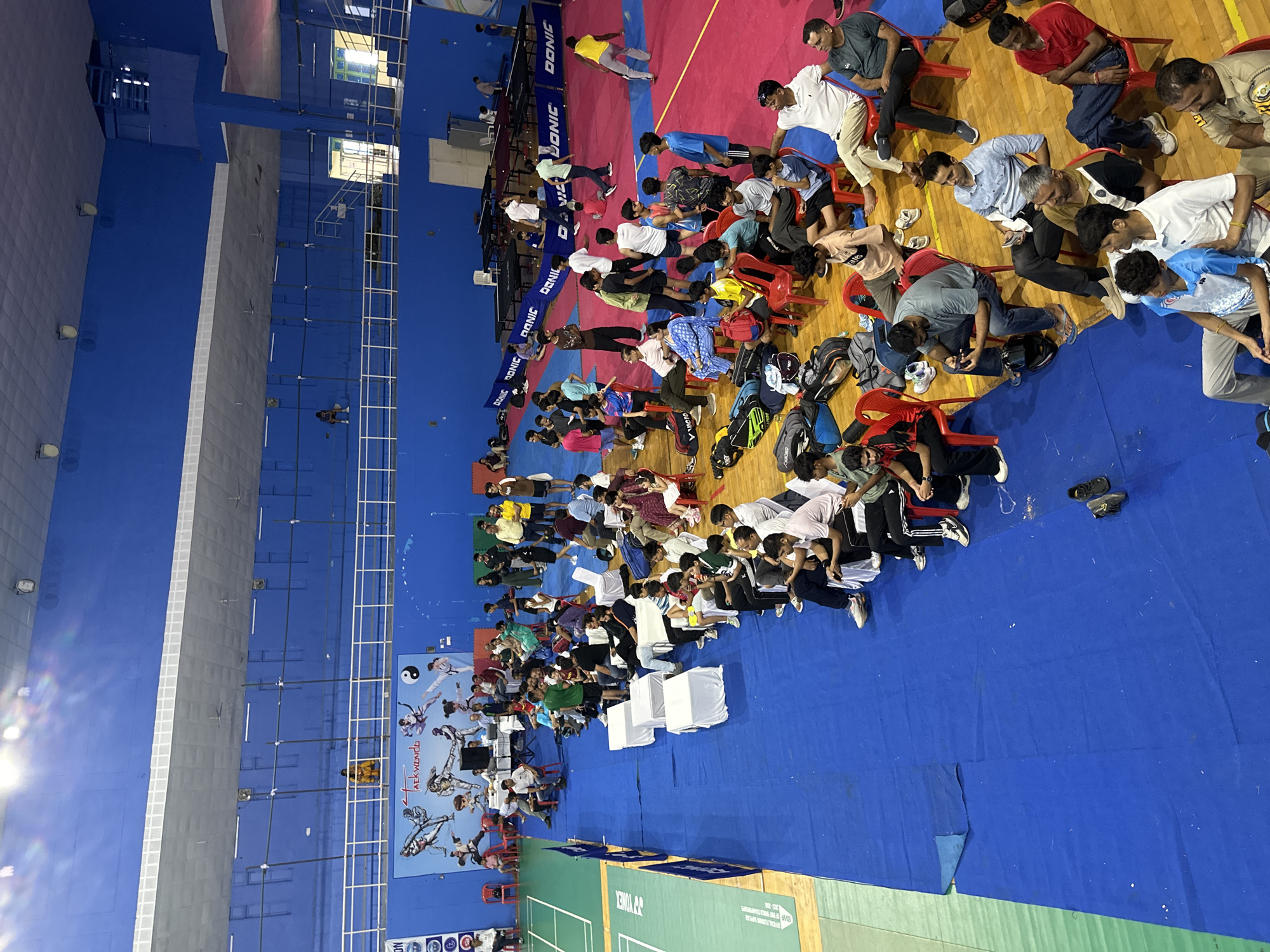 The image shows a wide, overhead view of an indoor sports arena, likely for taekwondo or a similar martial art. The arena features bright blue walls and a gray ceiling with visible lighting fixtures. A large banner depicting taekwondo athletes is displayed on the blue wall.
In the foreground, a blue mat covers the floor, bordered by a red competition area, likely for a ring or court. Several rows of chairs are set up, occupied by a diverse crowd of spectators, many of whom are dressed casually. Some individuals are looking at their phones, while others are engaged in conversation. There are also several sports bags and equipment scattered around the seating area.
Further back, the space transitions to a wooden floor with what appear to be several table tennis tables, indicated by the blue "DONIC" branded table covers. People are seen standing and sitting around these tables, some appearing to be participants or officials. The overall atmosphere suggests an ongoing sporting event or competition, with a mix of active participants and seated onlookers. The lighting appears to be a combination of natural light from high windows and artificial lighting from the ceiling. The location is not specified beyond being indoors.