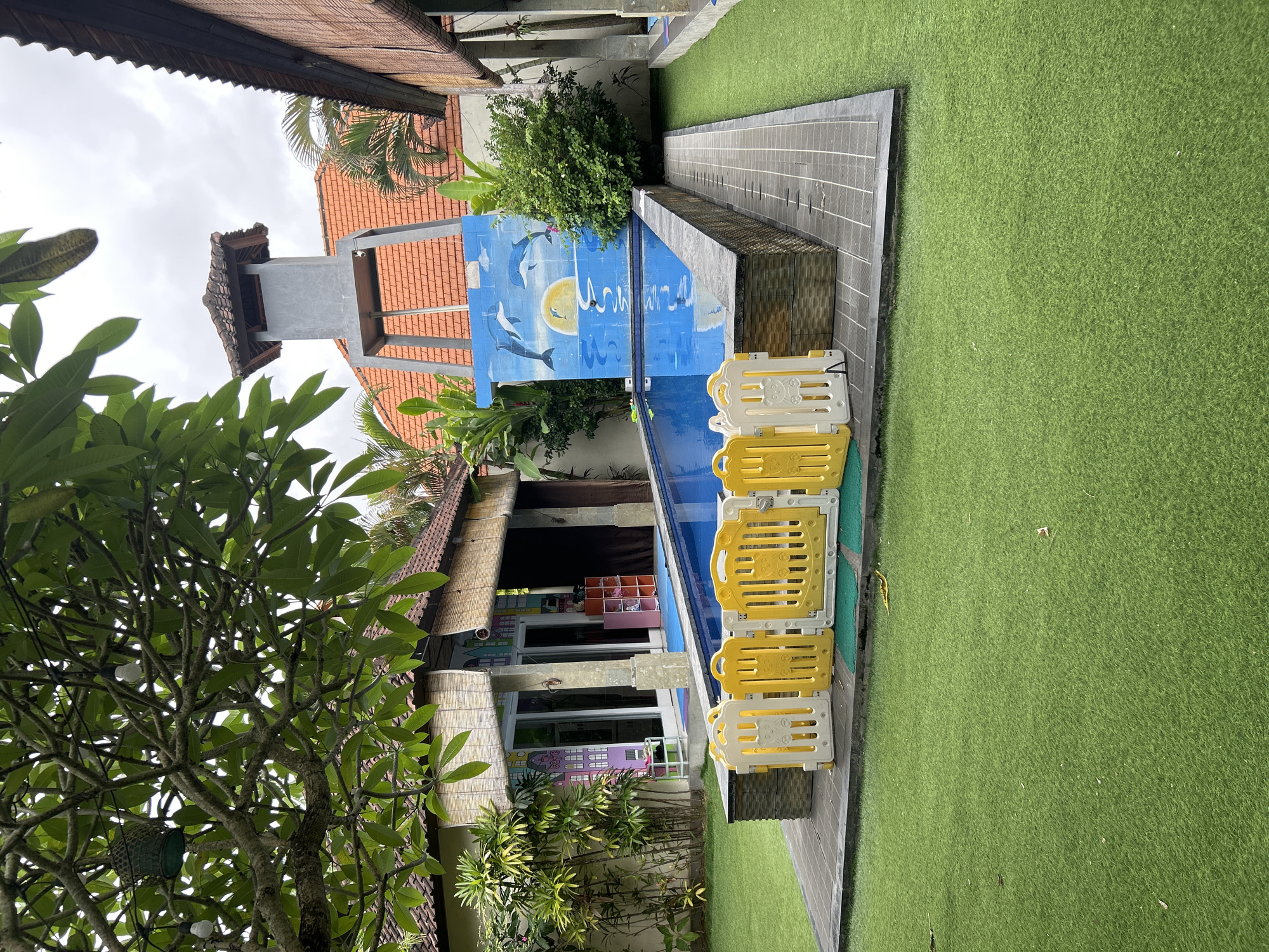 The image depicts an outdoor swimming pool area in Kuta, Indonesia. A rectangular swimming pool with bright blue water is centrally located, surrounded by a tiled deck. Yellow and white plastic safety fences are positioned around the edge of the pool. To the left of the pool is a building with a thatched roof and large glass doors, adorned with colorful murals. To the right, a building with bamboo blinds is visible, and a tower-like structure stands behind the pool, topped with a tiled roof. The ground is covered with artificial turf. The sky is overcast, and lush green foliage is present throughout the scene.