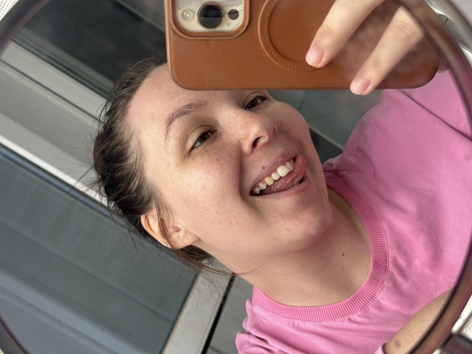A young woman is taking a selfie in a mirror. She is wearing a pink t-shirt and has her hair pulled back. She is sticking her tongue out and smiling. The background appears to be an indoor setting with vertical blinds visible through the mirror. The lighting suggests it is daytime.