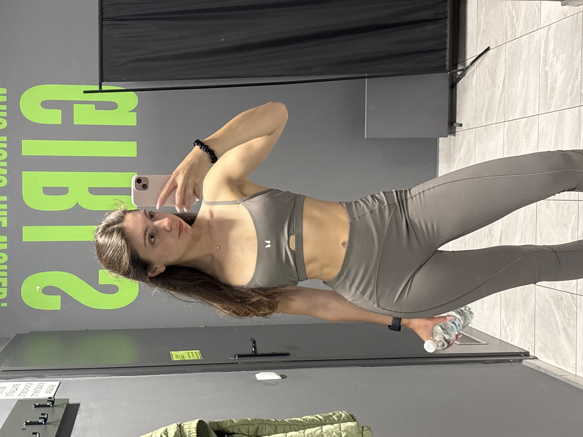 A young woman with long, dark brown hair is taking a mirror selfie in a locker room. She is wearing a grey sports bra and matching leggings. She is holding a water bottle in her left hand and her right hand is raised, holding her phone. The background of the image shows grey walls with some green lettering that appears to say "GIRLS". There is also a black curtain and a rack with hangers on the left side of the image. The floor is tiled with light-colored tiles. The woman is positioned in the center of the frame. The locker room is likely in a gym or fitness facility located in Varna, Bulgaria.