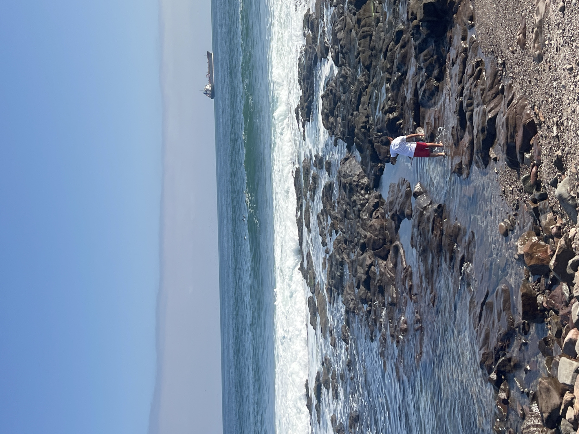 The image depicts a rocky beach on the coast of Arica, Chile, on a clear day. In the distance, a cargo ship is visible on the ocean. Closer to the shore, waves crash against dark, jagged rocks. A person wearing a white shirt and red shorts is standing barefoot in a shallow pool among the rocks. The transition from the rocky area to the beach is marked by smaller pebbles and stones.