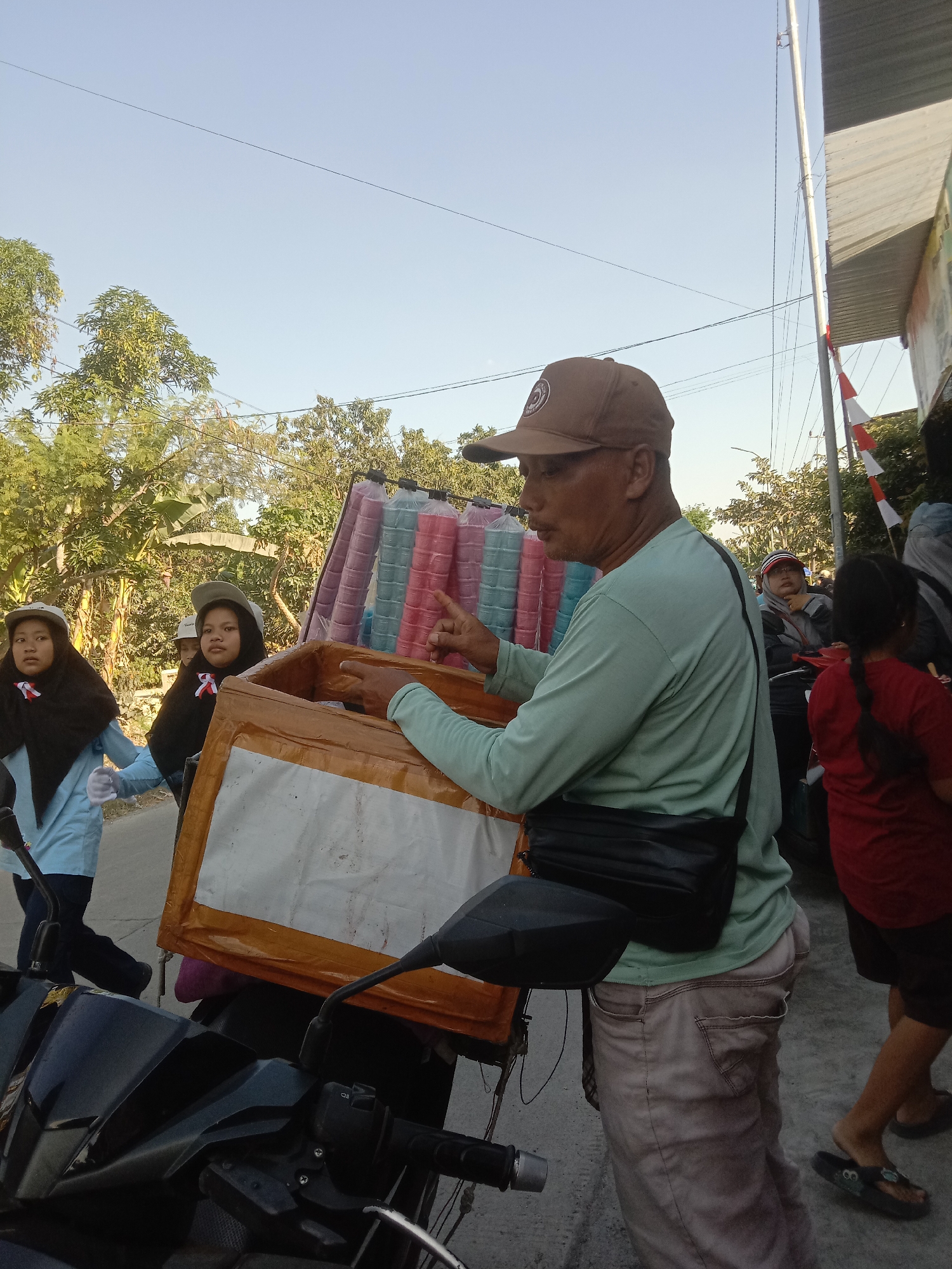 In Tulungrejo, Indonesia, a man wearing a light green long-sleeved shirt and a brown baseball cap is standing next to a motorcycle. He has a black bag slung across his chest. In front of him is a tan cooler box with a white label on its side. Behind him, a rack is attached to the motorcycle, filled with rows of colorful plastic cups containing ice cream, predominantly in shades of pink, blue, and purple.
To the left of the man, two young girls in light blue shirts and black hijabs, wearing white gloves, are walking. They are also wearing caps and have red, white, and blue ribbons pinned to their shirts. To the right of the man, another person in a red shirt and dark shorts is visible, and further back, a person wearing a red and white cap is seated on a motorcycle. The environment appears to be a street, with trees and buildings in the background, and overhead wires are visible against a clear sky. The scene suggests a street vendor preparing to sell ice cream.