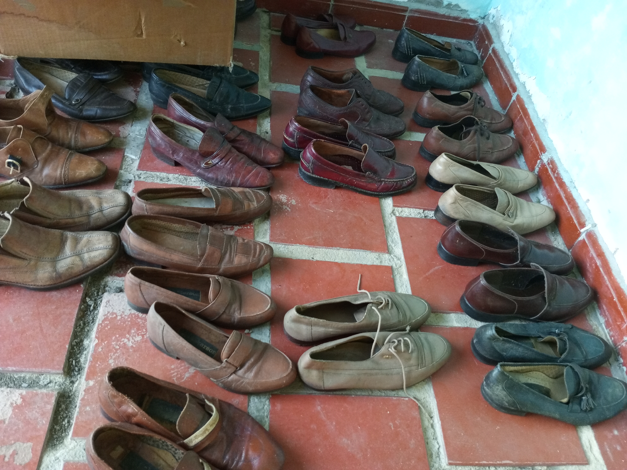 The image depicts a collection of used shoes arranged on a red brick floor. The shoes are mostly loafers and slip-ons, exhibiting a variety of colors including brown, black, tan, and red. The shoes appear worn, some with visible signs of age and wear. Some shoes are aligned in pairs while others are scattered. At the top left of the image, a cardboard box is partially visible. The scene is indoors, with a wall visible on the right side of the image. Based on the location provided, the image is taken in Palo Negro, Venezuela.
