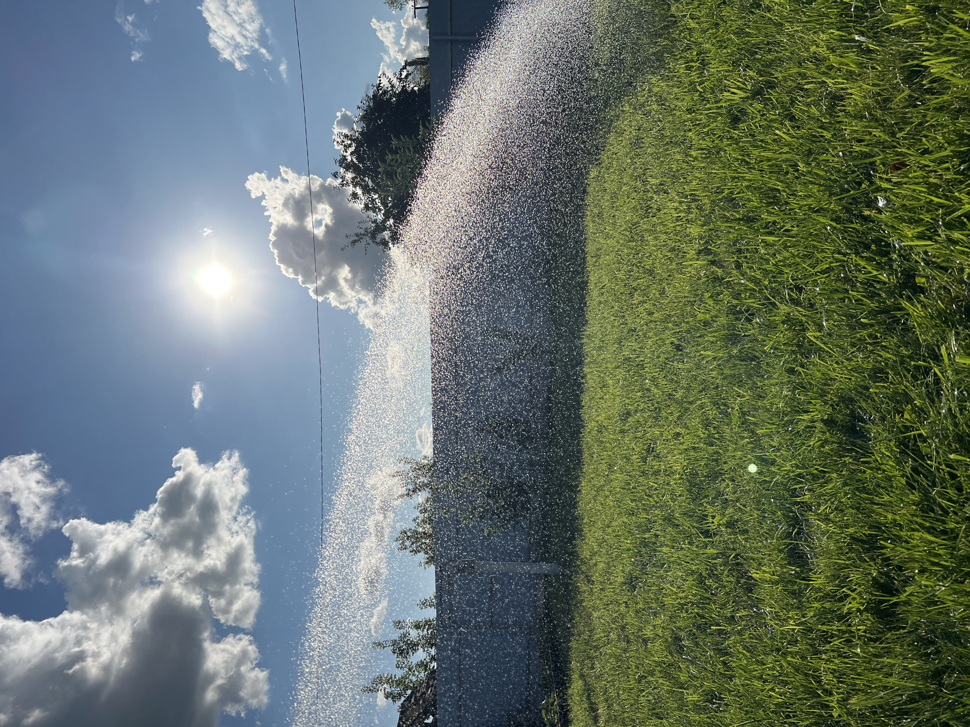 The image depicts a sun-drenched backyard scene with a lawn sprinkler in operation. Lush green grass covers the foreground, with water droplets from the sprinkler creating a shimmering curtain across the middle ground. Behind the grass, a grey fence runs horizontally, partially obscured by the spray. Trees and glimpses of rooftops are visible beyond the fence, suggesting a residential area. The sky is a vibrant blue, punctuated by scattered white clouds. The sun is high in the sky, casting a bright glare and lens flare onto the scene. The image was captured in the city of Salavat, Russia. There are no people or animals visible in the frame.
