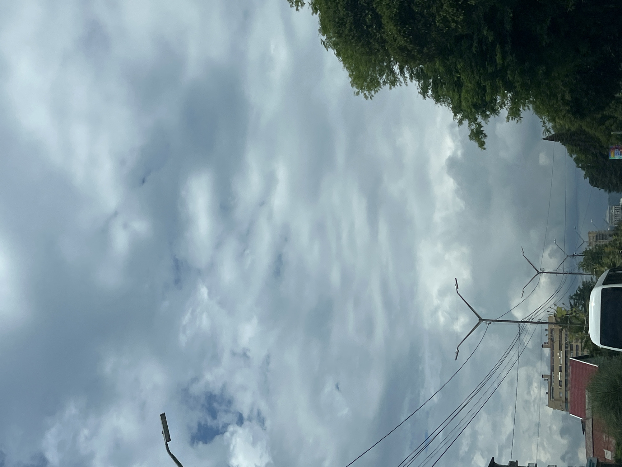 The image shows a cloudy sky over a city street. There are several streetlights visible, along with power lines. On the left side of the frame, there is a building with a red roof. A white bus is partially visible in the lower center of the image. On the right side of the frame, there are lush green trees. The overall impression is of an overcast day in an urban environment.