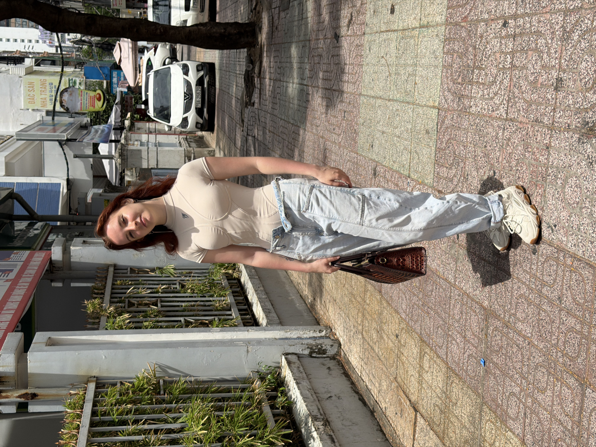 A young woman stands on a sidewalk in Nha Trang Ward, Vietnam. She faces the camera, holding a brown handbag, and is wearing a tan top and light blue jeans. The sidewalk is made of patterned red and beige tiles. Behind her is a low concrete wall covered in green plants. Further back, there are buildings and a white car parked on the street. A tree trunk is visible at the top of the frame.