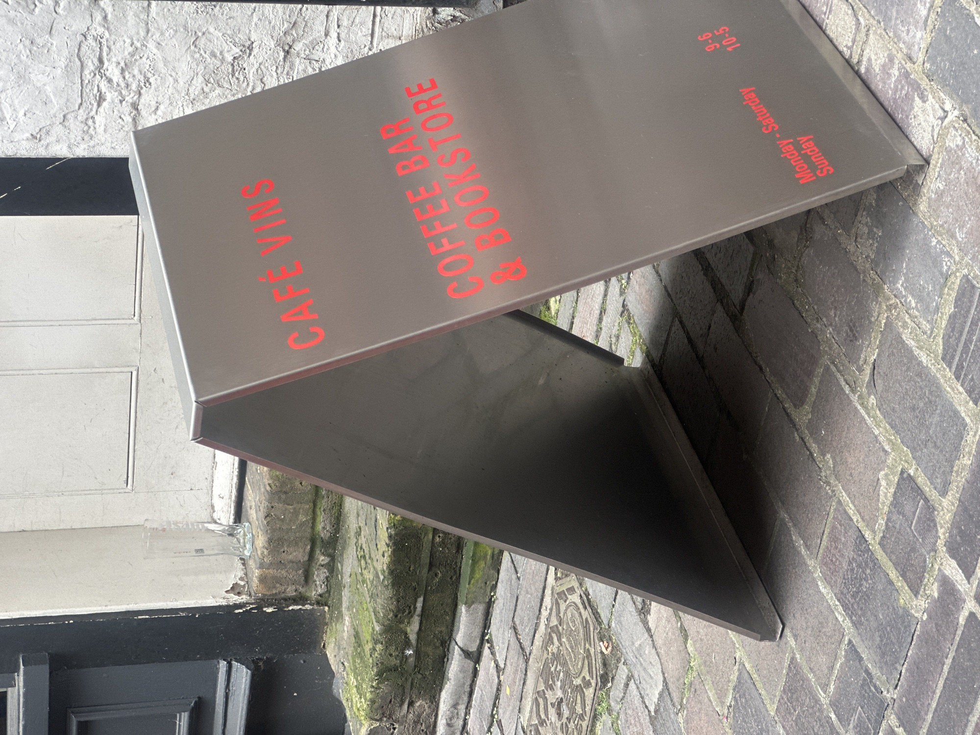 The image shows a metallic A-frame sign with red lettering on a cobblestone street. The sign is placed on a sidewalk next to a building with a stone facade and a dark-colored door on the left. The sign reads "CAFÉ VINS" at the top, and below that, "COFFEE BAR & BOOKSTORE". Further down, it lists operating hours: "Monday - Saturday 9-6" and "Sunday 10-5". The environment appears to be an outdoor setting in a city, with a drain cover visible on the cobblestones. The overall impression is one of an establishment advertising its services. The image was taken in London, United Kingdom.