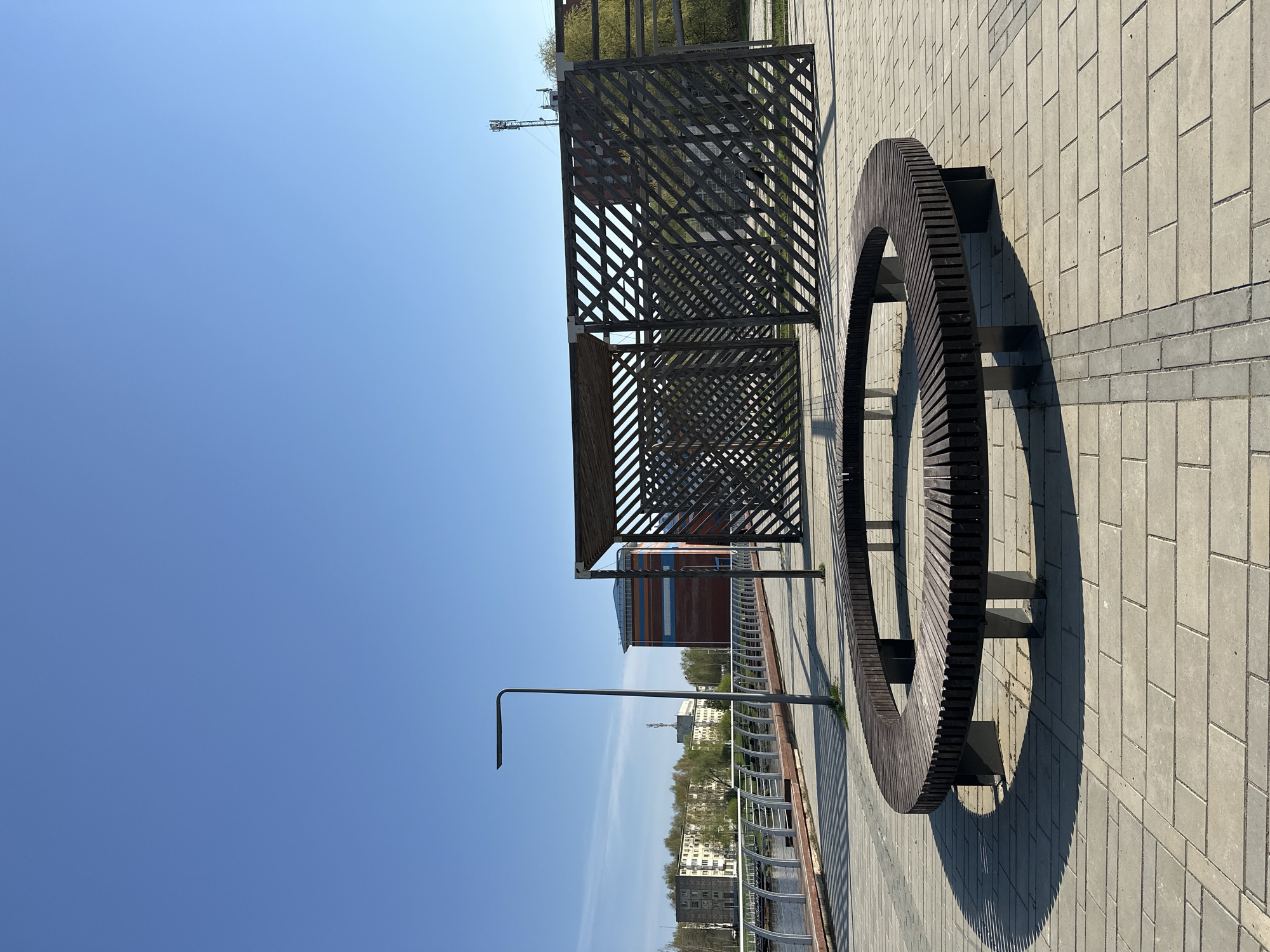 The image shows an outdoor scene on a paved area in Votkinsk, Russia. A semi-circular wooden bench with dark metal supports is positioned to the right. Next to it is a rectangular gazebo-like structure made of dark wood with a lattice design. A single lamppost is visible in the middle ground. In the background, there is a view of buildings, trees, and a waterway with a metal railing along the edge. The sky is clear blue. The scene appears to be taken during daytime with sunlight casting shadows from the structures.