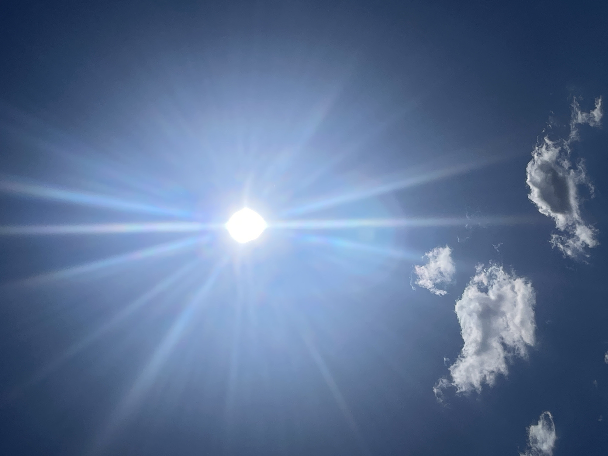 A bright, daytime scene captures the sun prominently positioned in a pale blue sky. Radiating outward from the sun are vibrant, intense rays of light. Several white, fluffy clouds are visible against the blue backdrop, situated to the right of the sun. The location is Unknown, Unknown.