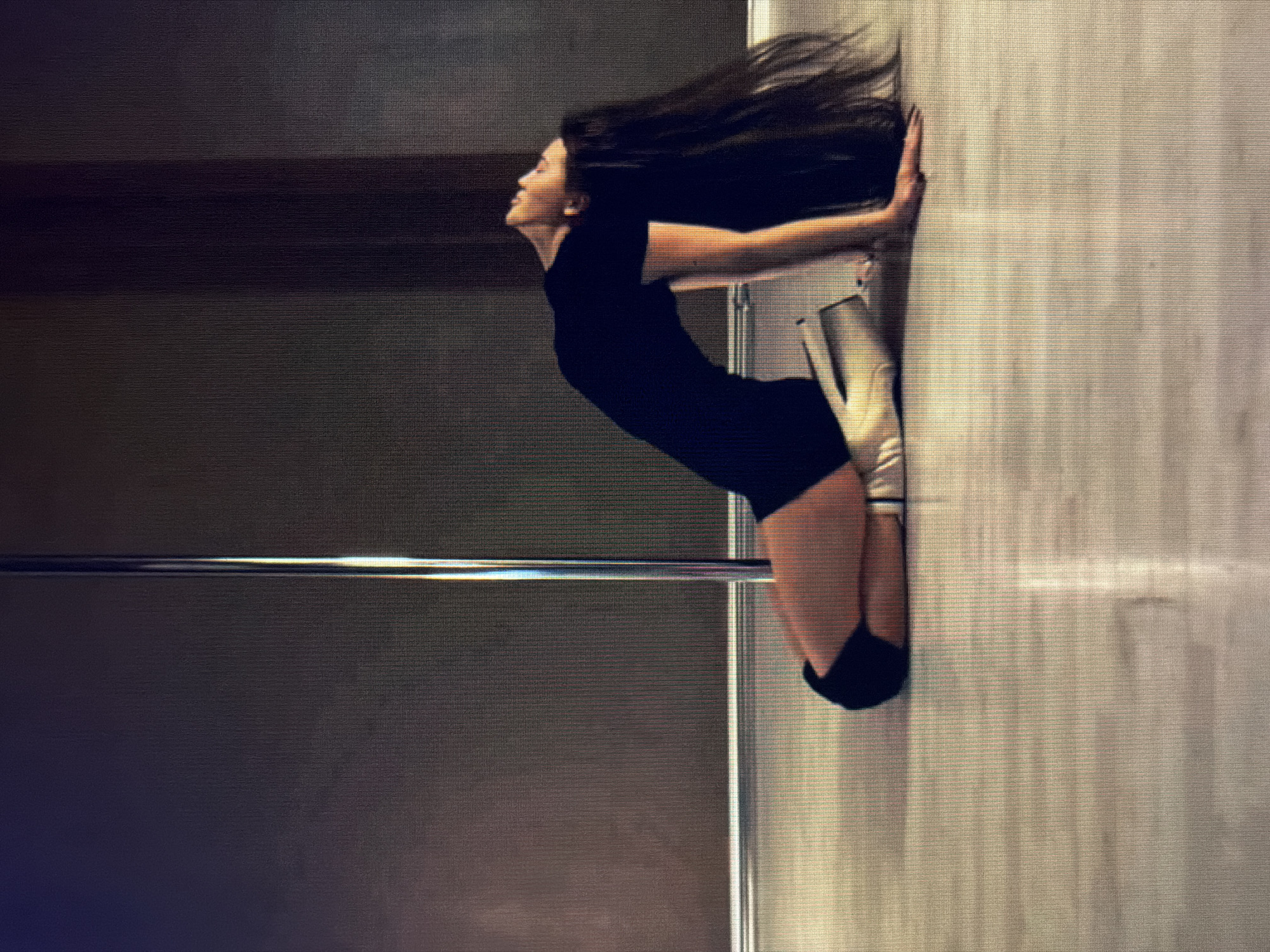 A person is kneeling on a polished wooden floor with their back arched and head thrown back. They are wearing a black leotard and black knee pads. Their long, dark hair is flowing behind them. A metal pole stands to their left, and the background is a dark, textured wall. The scene appears to be indoors, possibly a dance studio or gym. There is no identifiable information about the city of Sirius, Russia, within the image.