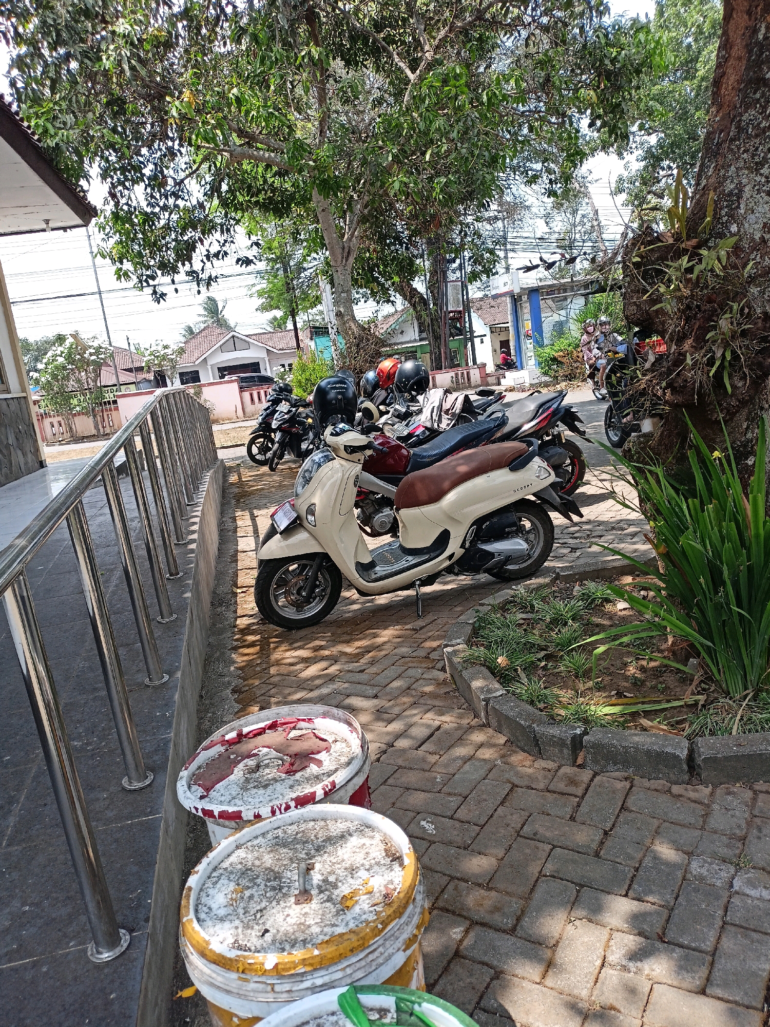 A bright, sunny day in Kewaluhan, Indonesia, is captured in this photograph. In the foreground, two large, weathered buckets sit on a herringbone-patterned brick walkway. The buckets appear to have been used for paint or some other material, with remnants of red and yellow paint visible on their rims and surfaces. A stainless steel railing runs along the left side of the frame, bordering a concrete structure.
Beyond the railing, a beige scooter with a brown seat is parked on the brick path. Several other motorcycles, including a red and black one and a black one with a distinctive red stripe, are parked behind it. The motorcycles are also adorned with helmets. In the background, typical Indonesian houses with tiled roofs are visible, interspersed with trees that provide shade. Power lines crisscross the sky, a common sight in urban and suburban areas. Further back, a group of people on a motorcycle, including a child, are seen approaching, their presence adding a sense of movement and life to the scene. The overall impression is that of a quiet, everyday scene in a residential area in Kewaluhan.