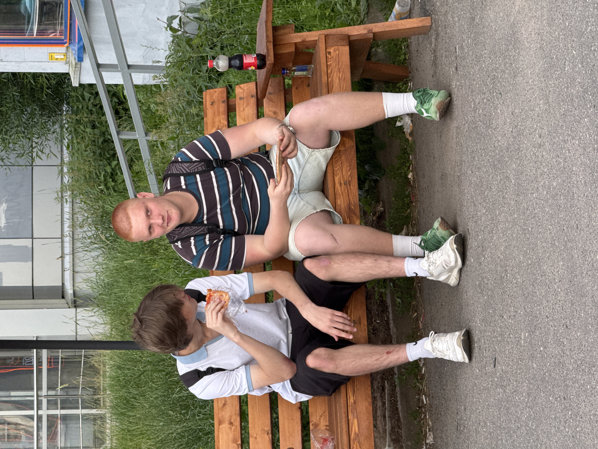 Two young men are sitting on a wooden bench outdoors, with grass and a paved surface around them. The man on the left has red hair and is wearing a striped t-shirt and light-colored shorts. He is holding a small, rectangular object, possibly a phone, in his hands. The man on the right has brown hair and is wearing a white t-shirt with blue trim and black shorts. He is eating a slice of pizza, which is wrapped in plastic.
Several drink bottles are visible on a wooden table attached to the bench. One is a clear bottle with a red cap, and another appears to be a bottle of soda. There is also a small blue bottle that might contain alcohol. The ground around the bench is a mix of grass and asphalt. In the background, part of a building with windows is visible. The lighting suggests it is daytime, and the presence of green grass and trees indicates fair weather. The men appear relaxed and are engaged in eating and holding their devices. There is no visible text in the image.