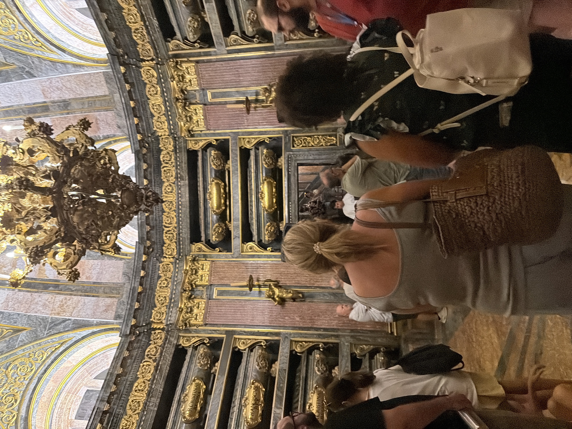 The image depicts the Royal Pantheon within the Monastery of San Lorenzo de El Escorial, located in San Lorenzo de El Escorial, Spain. A group of people are observing the ornate mausoleums lining the circular walls of the pantheon. The architecture is characterized by rich gold detailing against dark marble and pink columns. A large, elaborate chandelier hangs from the domed ceiling. Several individuals are visible from behind, some carrying bags and backpacks. The scene is one of respectful observation within a historically significant burial site.