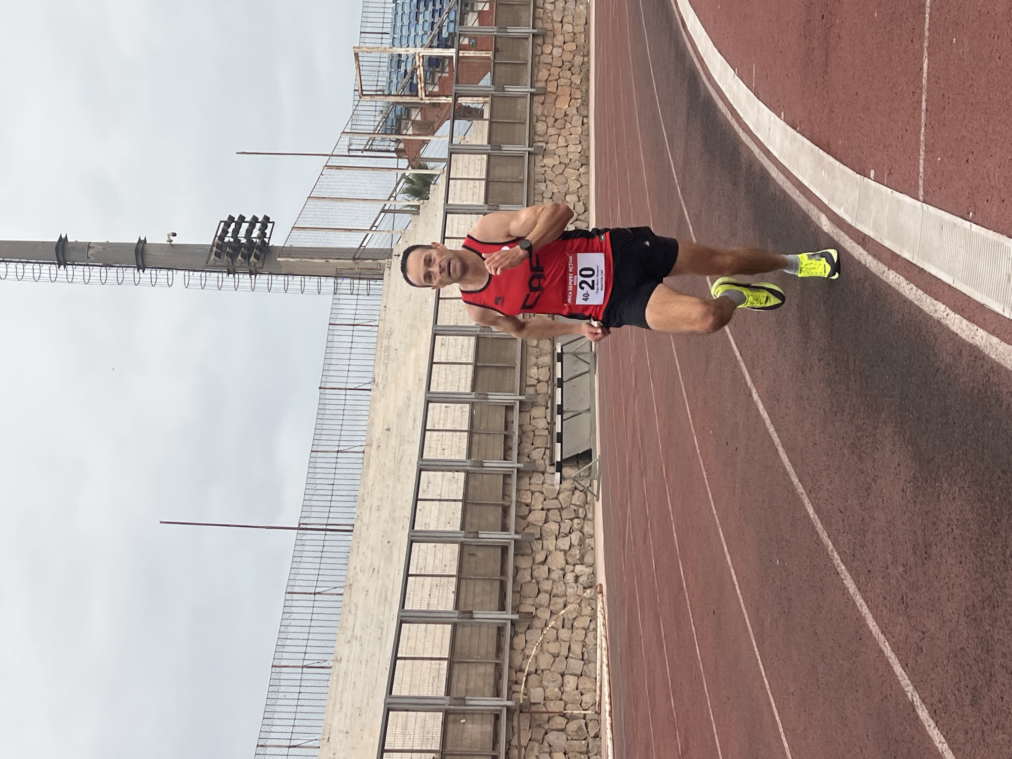 A man is running on a red athletic track at an outdoor stadium. He is wearing a red tank top with a bib number "40-20", black shorts, and yellow running shoes. The stadium has concrete bleachers in the background, a stone wall, and a tall light tower with lights and a ladder. The sky is overcast. The image appears to be taken from a slightly low angle, focusing on the runner. The location is identified as the Estadio Municipal de Arica, located in Arica, Chile, a coastal city in the far north of the country.