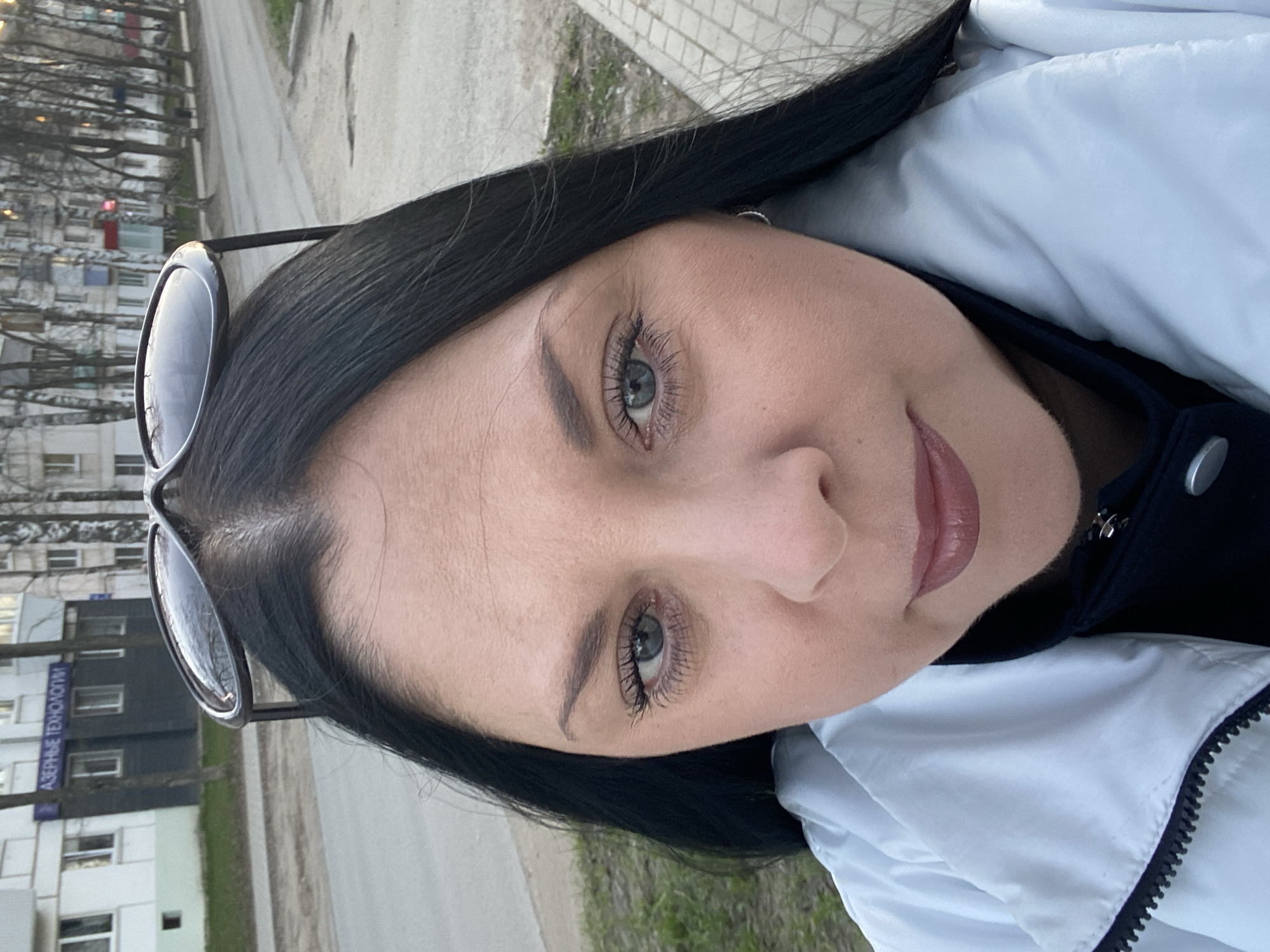 A close-up, rotated selfie shows a woman with dark hair and light eyes, wearing a white jacket. Sunglasses are perched on top of her head. The background reveals a street and buildings, suggesting an urban setting. A building with the inscription "АЗЕРНЫЕ ТЕХНОЛОГИИ" in Russian is visible. This places the location in Chaikovsky, Russia, as "АЗЕРНЫЕ ТЕХНОЛОГИИ" likely refers to the company "Azerneft Technology", which has a branch in that city. The scene suggests an outdoor setting during daylight.