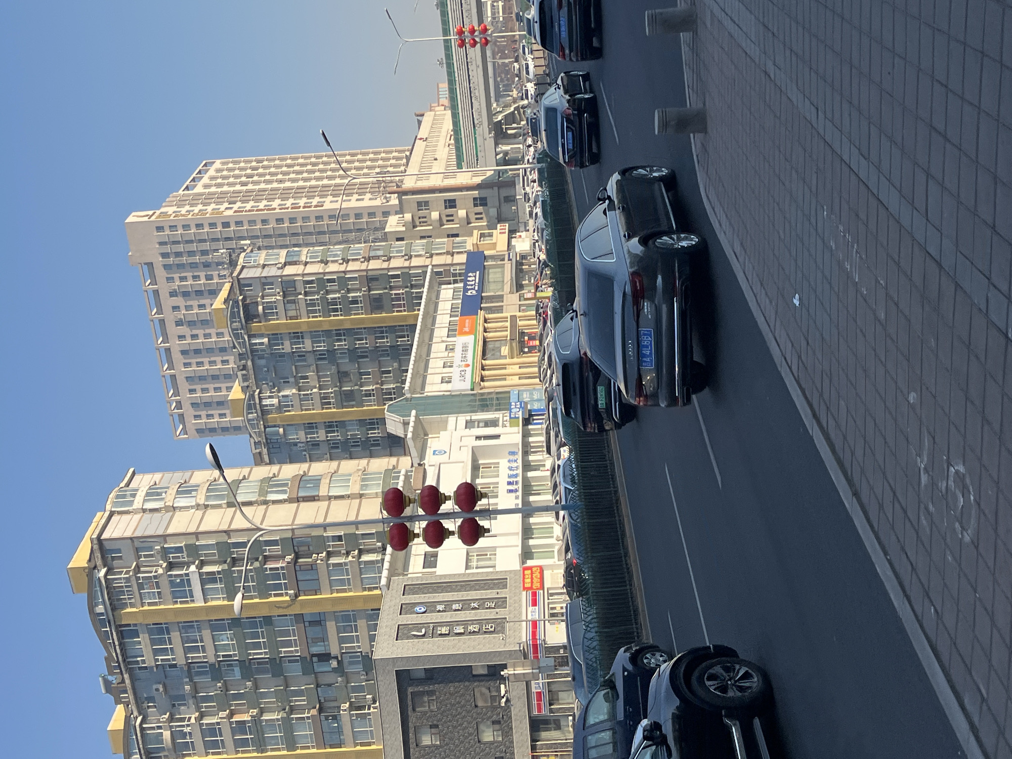 The image shows a street scene in Changchun City, China, with buildings, cars, and street furniture. The cars are lined up on the road, with some parked along the side of the street. A red light fixture is visible. The buildings appear to be high-rise apartments or commercial structures, with one featuring signage that includes Chinese characters for "Lefeng Hotel" (麗枫酒店). Additional signs include "禅意沐足" and "旺铺出租" (retail space for rent) with a phone number, "13019128429." Further down the street, there is a sign indicating "吉林农商行" (Jilin Rural Commercial Bank) with the letters "JLRCB." The visible license plates on the cars read "吉A4L887" and "BA F24562." The sky is clear and blue, suggesting a sunny day.