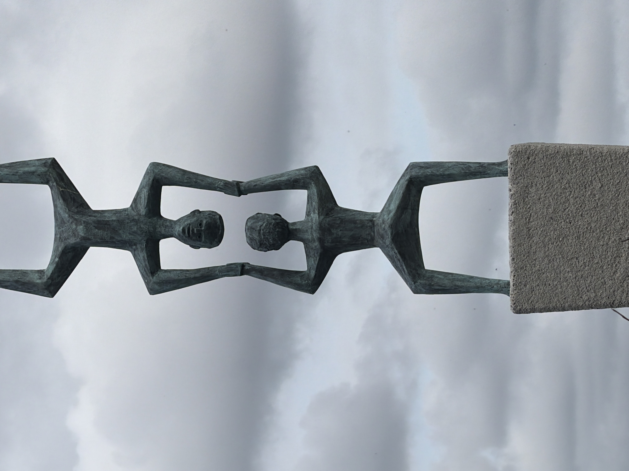 The image shows a bronze statue of three stylized human figures stacked vertically on a stone pedestal. The bottom figure is kneeling, supporting the middle figure who is standing and holding the top figure upside down. The top figure's head is resting on the middle figure's hands, and its legs are extended upwards. The sculpture is positioned against a backdrop of a cloudy sky. This sculpture is titled "The Three Graces" and is located in the central plaza of the city of Rijswijk, Netherlands.