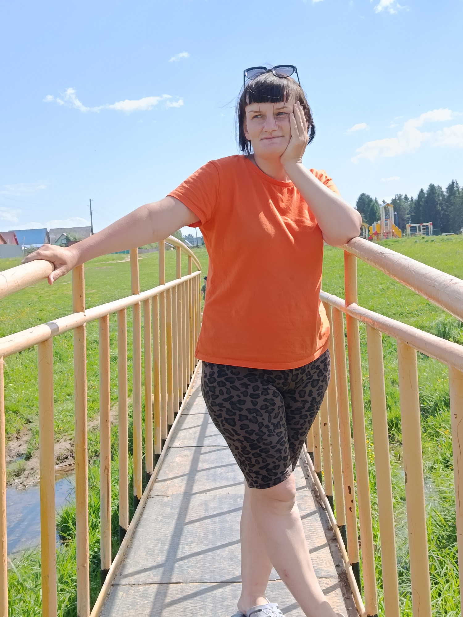A woman stands on a bridge in an outdoor setting. She is wearing sunglasses on her head, an orange t-shirt, and patterned shorts. Her left hand rests on her face while her right arm is extended, holding onto the yellow railing of the bridge. The bridge has a metal deck with railings. Below the bridge, there is vegetation and what appears to be a small stream or body of water. In the background, there is green grass, trees, and a playground structure. In the distance, a structure is visible on the left side of the frame. The sky is blue with scattered clouds. The location is unknown.