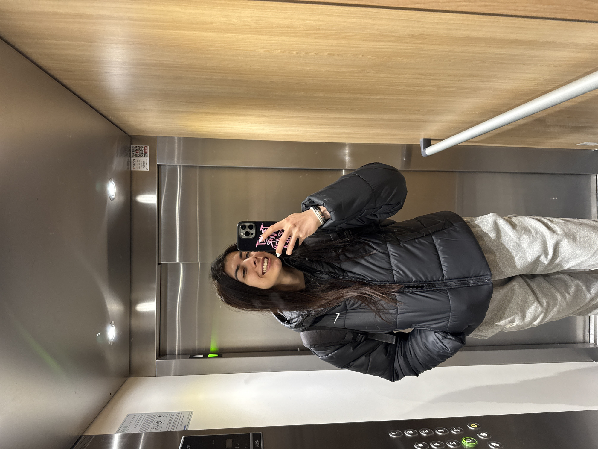 A young woman stands in an elevator, taking a selfie. She is wearing a black puffer jacket and gray sweatpants. Her phone case has pink writing. The elevator's interior features stainless steel walls and a wooden ceiling. The elevator control panel is visible, along with a grab bar. The location is an elevator within a building in Sofia, Bulgaria; however, the specific building cannot be determined from this image alone.
