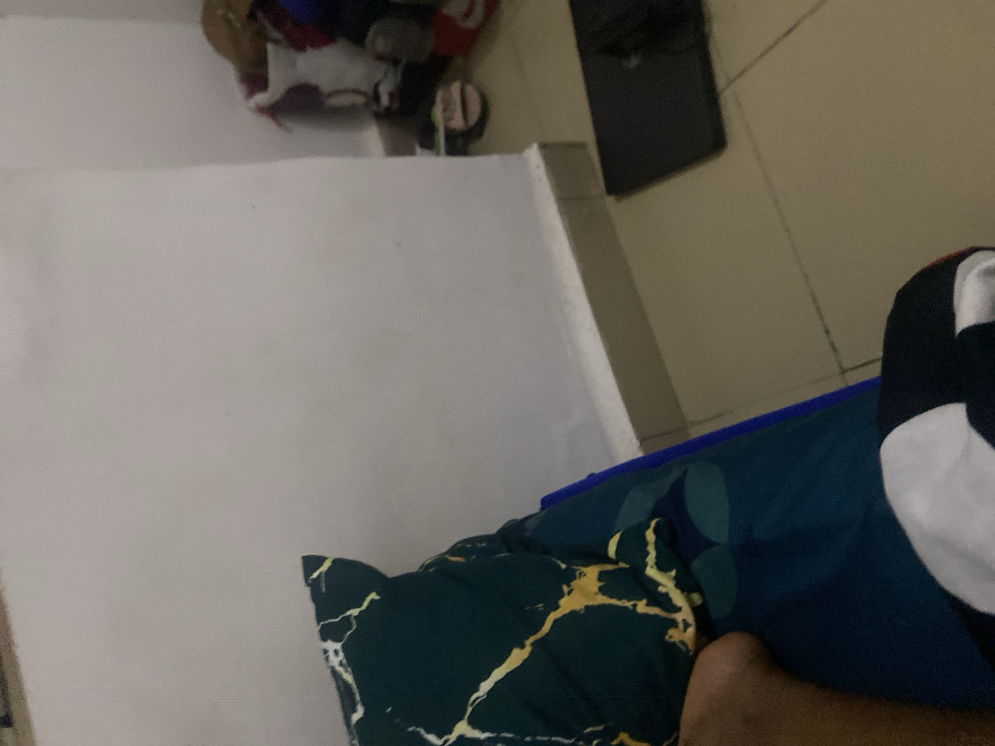 The image shows a close-up of a bed with a pillow that has a dark green and yellow marble pattern. To the right of the pillow, there is a glimpse of a person's leg. The bed is covered with a blue sheet that has a subtle circular pattern. In the background, a white wall meets the floor, which is tiled. On the floor, there are several pairs of shoes, including one pair of brown sandals, and a black laptop. The overall scene appears to be a bedroom environment. The location is identified as Howayi, Nigeria.