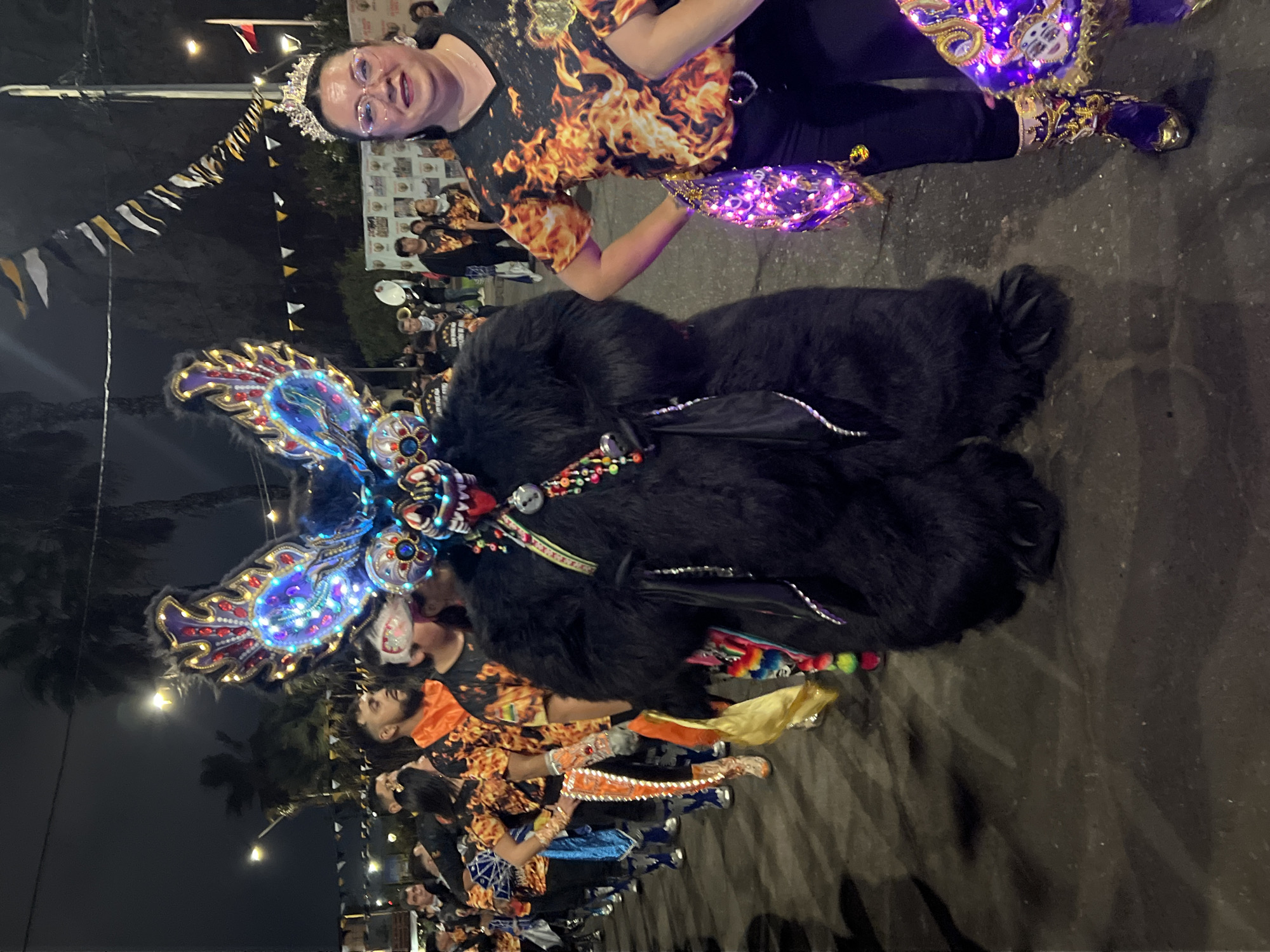 The image depicts a nighttime street parade in Arica, Chile, likely part of a cultural festival. In the foreground, a person in an elaborate, large, black furry costume with a colorful, illuminated mask resembling a dragon or mythical creature is the central figure. The mask has prominent, iridescent blue and red wings with intricate patterns. A woman wearing a crown and glasses, dressed in a black shirt with a flame design, stands to the right of the costumed figure. She is holding a purple, illuminated scarf or sash. Behind them, a procession of people in matching orange and black flame-patterned shirts are visible, some carrying colorful sashes or banners. The street is paved, and overhead, festive flags and lights are strung across the scene. Palm trees are faintly visible in the dark background, and streetlights illuminate parts of the parade route. The overall atmosphere suggests a lively celebration.