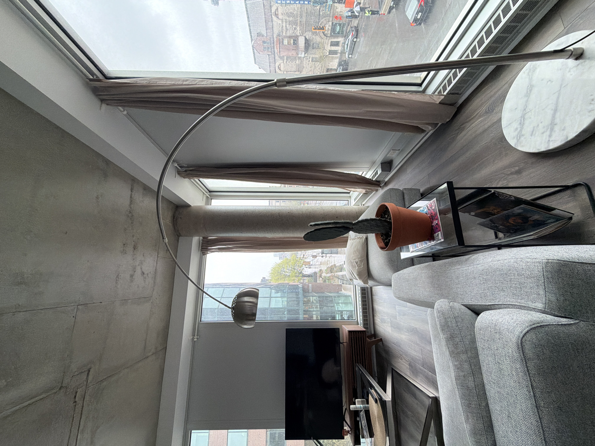 The image shows an interior view of a room with a focus on furnishings, decor, and a cityscape seen through the windows. The room includes a gray sofa, a black metal side table with books, and a cactus in a terracotta pot placed on the table. A floor lamp with a curved arm is present, along with a small round marble table. The windows are covered with beige fabric curtains, and a television is visible against a wall.
Outside the windows, a city view is visible, particularly from the top window which features a cityscape with brick buildings, including the Église du Gesù, and a glimpse of street traffic. A secondary window shows a more residential area with trees and lower buildings.
Based on the buildings visible, particularly the Église du Gesù, the location is likely in or around downtown Montreal, Canada.