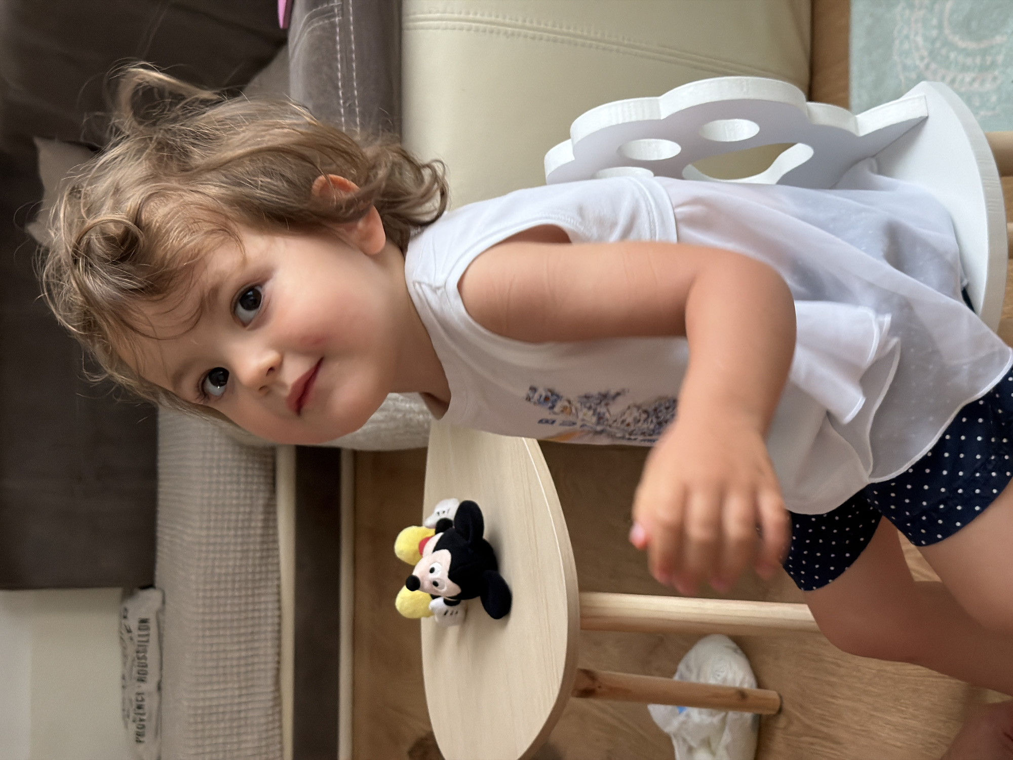 A young Caucasian female child with curly brown hair, brown eyes, and fair skin is seated on a white child's chair with a decorative, cut-out back. She is wearing a white sleeveless top and dark blue polka dot shorts. Her right arm is bent at the elbow, and her hand is slightly blurred due to motion. In front of her is a small, light-colored wooden table with rounded edges. On the table, a Mickey Mouse plush toy lies on its back. The background shows a living room area with a sofa and a rug. The image appears to be taken indoors, likely in a residential setting within Plovdiv, Bulgaria, although no specific landmarks or street views are visible to confirm this location.